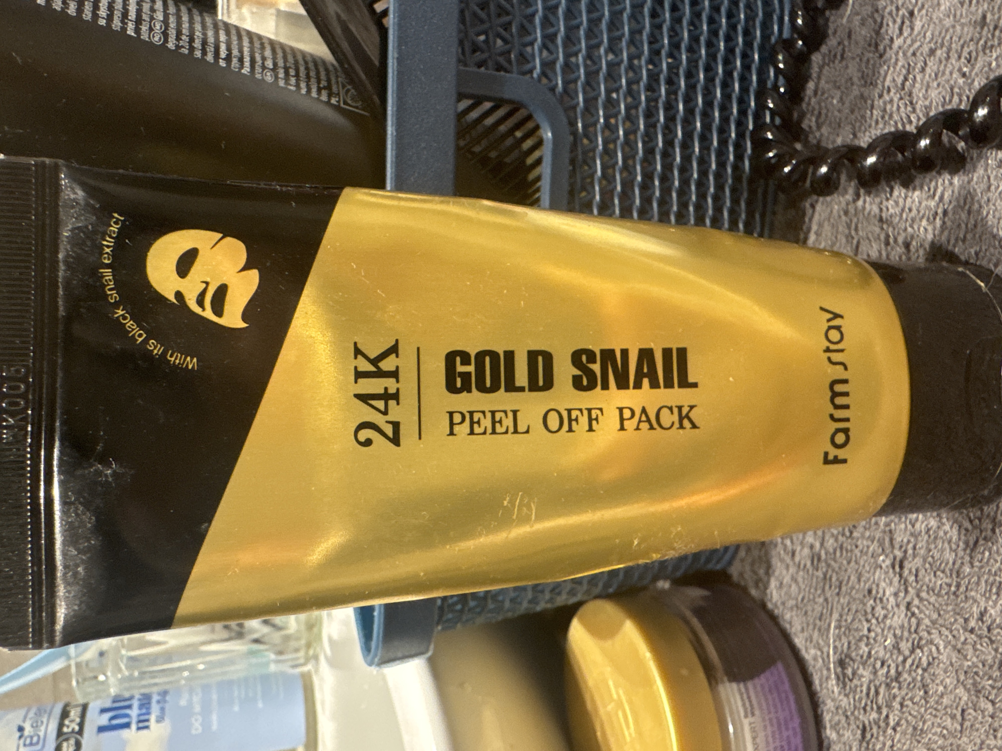A close-up, eye-level shot shows a tube of "24K Gold Snail Peel Off Pack" by Farm Stay, oriented vertically. The tube is primarily gold and black, with prominent text indicating its contents. The background is a blurry mix of what appears to be bathroom or vanity items, including a blue mesh holder and a black beaded cord. The lighting suggests an indoor setting, possibly a bathroom, during the day. There are no people visible in the image. The focus is entirely on the product packaging, highlighting its branding and product name. Visible text includes "24K GOLD SNAIL PEEL OFF PACK", "With its black snail extract", and "Farm stay". Additional text in various languages, likely ingredients or instructions, is present on the product and in the background but is mostly illegible. The scene appears to be a product display or a personal collection of cosmetic items.