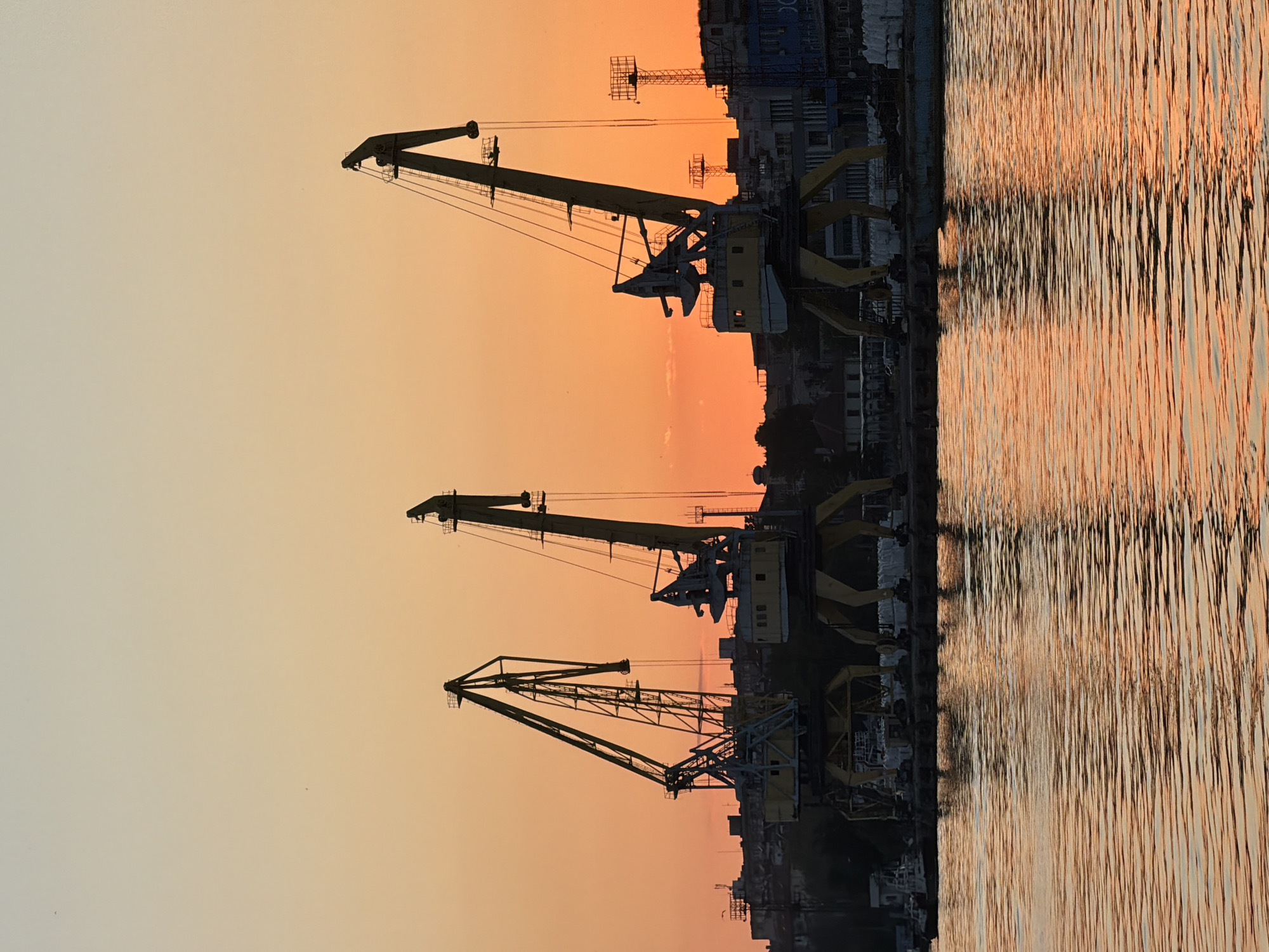 The image displays a row of three large harbor cranes positioned along a waterfront at sunset. The sky transitions from a pale yellow to a deep orange. The cranes are yellow with black silhouettes and stand against the skyline of Burgas, Bulgaria. Their reflections are visible in the water, appearing as vertical streaks of orange and gray. The cranes are located in an industrial area.