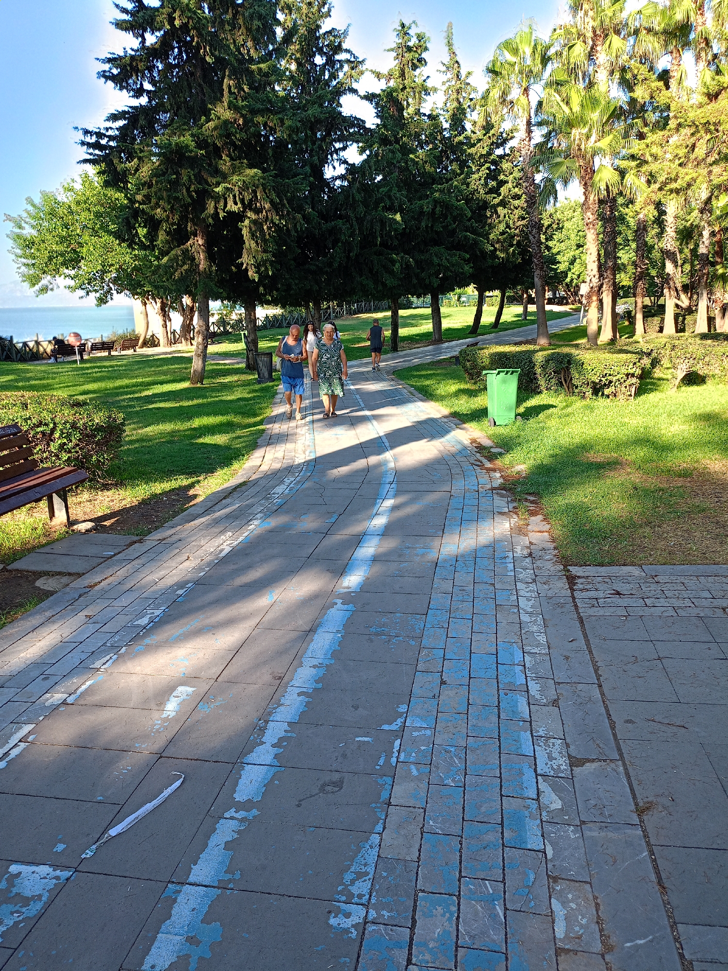 The image depicts a paved pathway in a park-like setting, likely in Antalya, Turkey, given the presence of palm trees and lush greenery. Two women are walking on the path, dressed in casual summer attire. The path is made of rectangular paving stones, with blue markings painted intermittently along its surface. The scene is bathed in sunlight, casting shadows from the trees onto the pathway. In the background, there are more trees, including tall evergreen trees and palm trees, a grassy area, and glimpses of what appears to be the sea or a large body of water beyond a wooden fence. A green trash bin is visible on the right side of the path, and a wooden bench is situated on the left. The overall atmosphere is serene and suggests a public recreational area.
