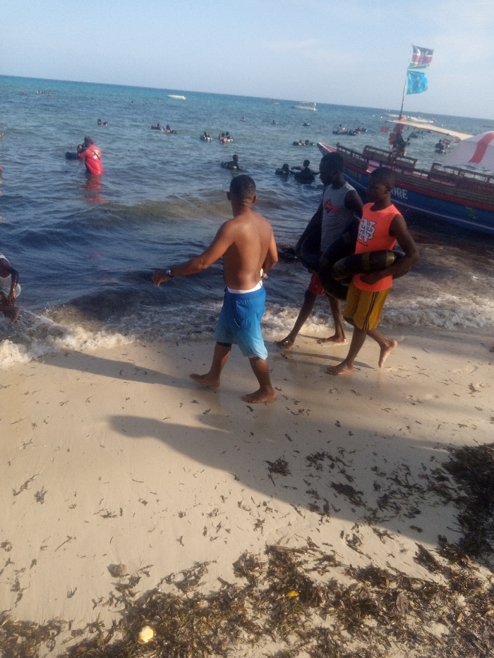 The image depicts a crowded beach scene in Shanzu ward, Kenya, under clear daytime conditions. In the foreground, three males walk barefoot along the sandy shoreline, moving from right to left. The leftmost individual is shirtless, wearing blue shorts, with his right arm extended towards the water. The middle individual wears a grey t-shirt and dark shorts, carrying a black inner tube. The rightmost individual wears an orange t-shirt and yellow shorts, also carrying a black inner tube.
The immediate sandy area at the waterline shows wet patches and small waves breaking, with scattered dark seaweed and debris. Beyond the individuals, the Indian Ocean water extends with numerous people swimming or wading. Many of these individuals are utilizing black inner tubes or similar floats. One person in the midground is distinguishable by a red shirt while partially submerged.
To the right, a large, blue wooden boat with a red and white stripe along its hull is visible. The name "SHANZORE" is stenciled on the boat's side. Two flags, one identified as the Kenyan national flag and another blue flag, are flown from a pole on the boat. A white canopy or tent-like structure is also visible on the boat. The sky above is clear and blue, indicating bright sunlight, which casts distinct shadows from the people onto the sand.
