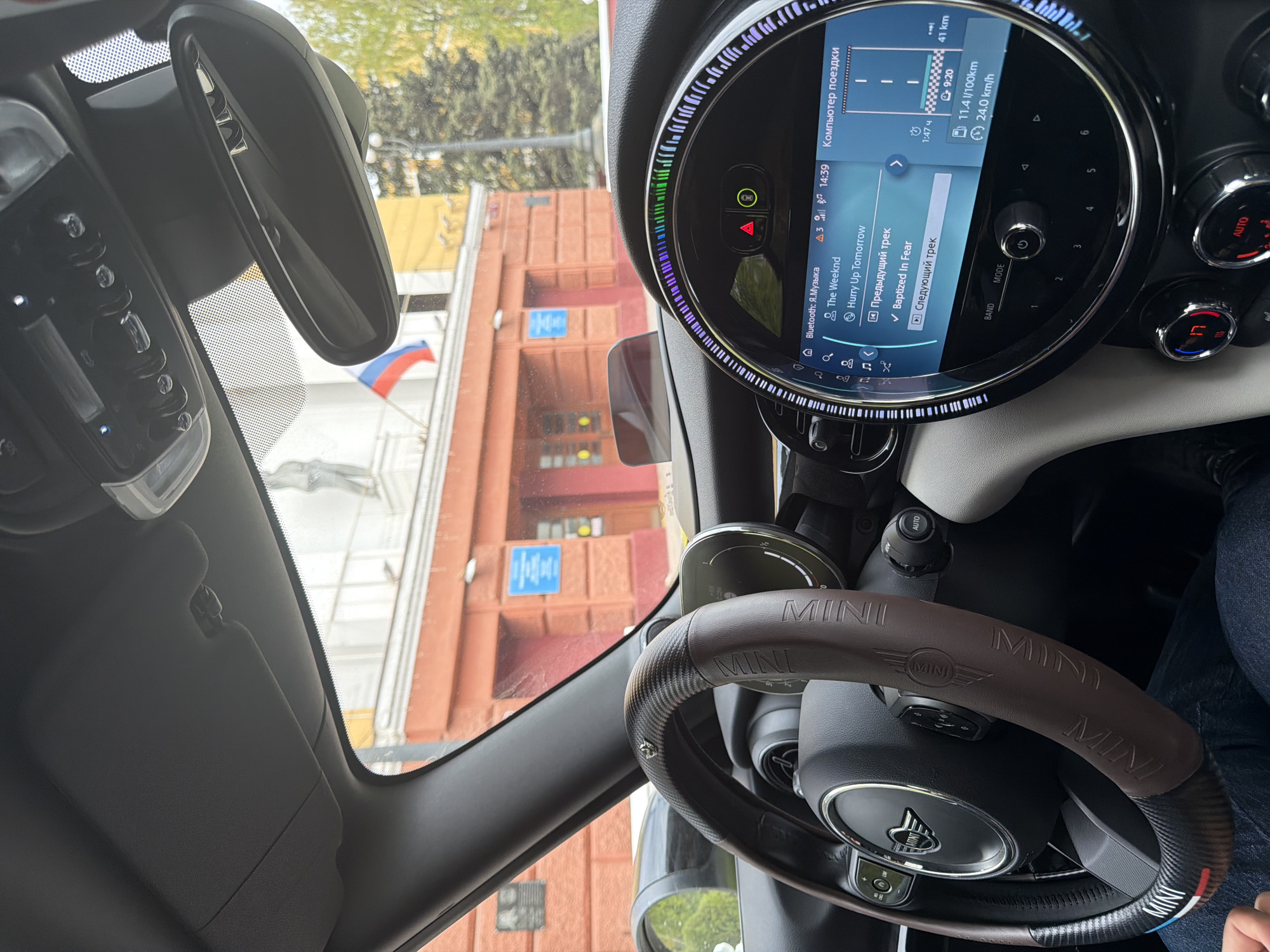 The image is taken from inside a Mini Cooper car. Through the windshield, a building is visible, identified by signage and the Russian flag as being in Krasnoyarsk, Russia. The dashboard of the car shows the stereo playing "Baptized In Fear" by The Weeknd, and trip information showing a distance of 41 kilometers, an average speed of 24 km/h, and fuel consumption of 11.4 l/100km. The steering wheel is brown and features "MINI" branding. A person is seated in the car, visible in the lower right portion of the frame.