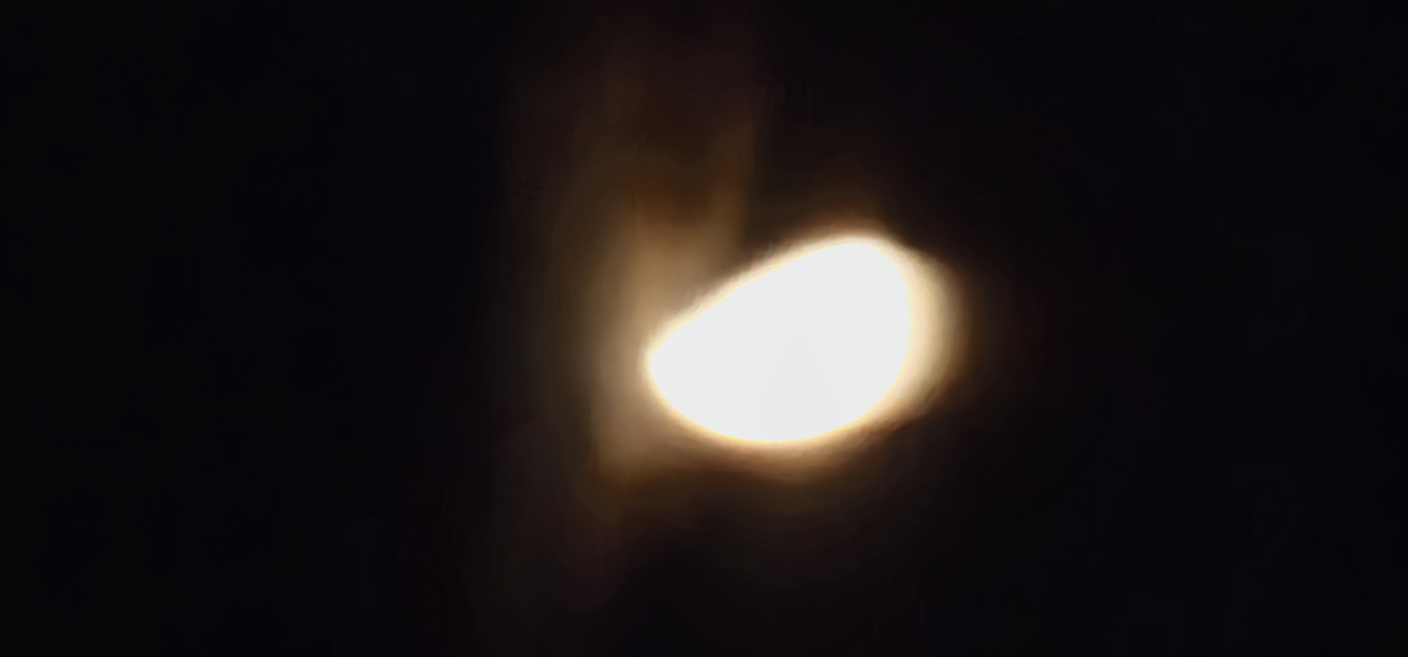 The image is a blurred, low-light photograph of a bright, luminous object against a dark background. The object appears to be roughly elliptical and emits a soft, warm glow, with a brighter, almost white center that fades to a yellowish-orange at the edges. There are subtle, wispy dark streaks or smudges around the object, suggesting it might be partially obscured by smoke, fog, or a similar atmospheric phenomenon. The overall impression is of a celestial body, perhaps the moon, or a distant, intense light source. There is no discernible location within the city of Villa Latina, Italy, or any identifiable people, subjects, objects, actions, or interactions. The image is abstract and lacks specific contextual details.