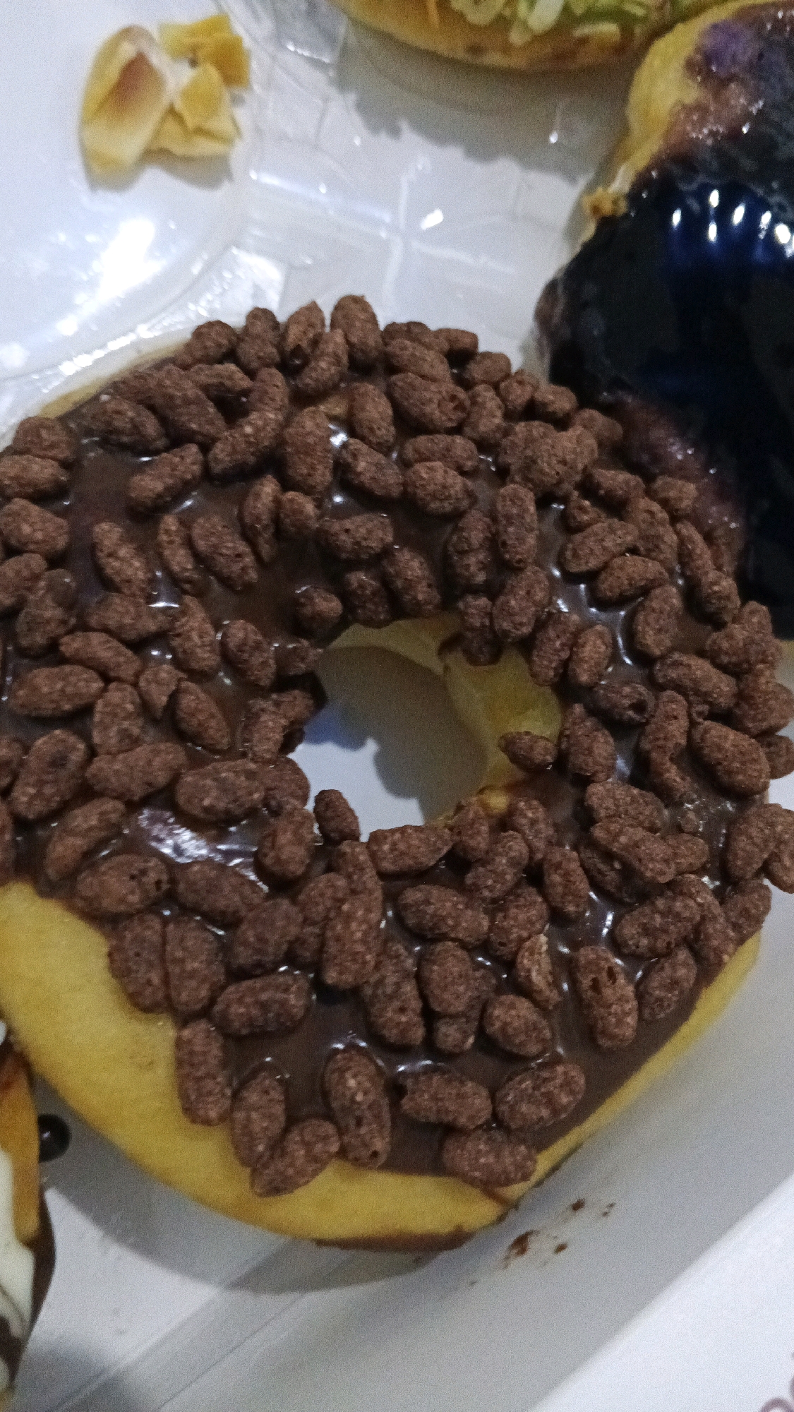 The image shows a close-up of a chocolate-frosted donut topped with chocolate cereal pieces. The donut is yellow and has a hole in the center. It is placed in a white box, suggesting it is a take-away item. In the background, partially visible, are other food items, including what appears to be another donut with dark frosting and some pieces of fruit or pastry. The lighting is artificial and the image quality is moderately low, with some graininess and blur. The scene is likely an indoor setting, possibly a bakery or a food stall in Semarang, Indonesia, given the provided context. No people are visible in the image.