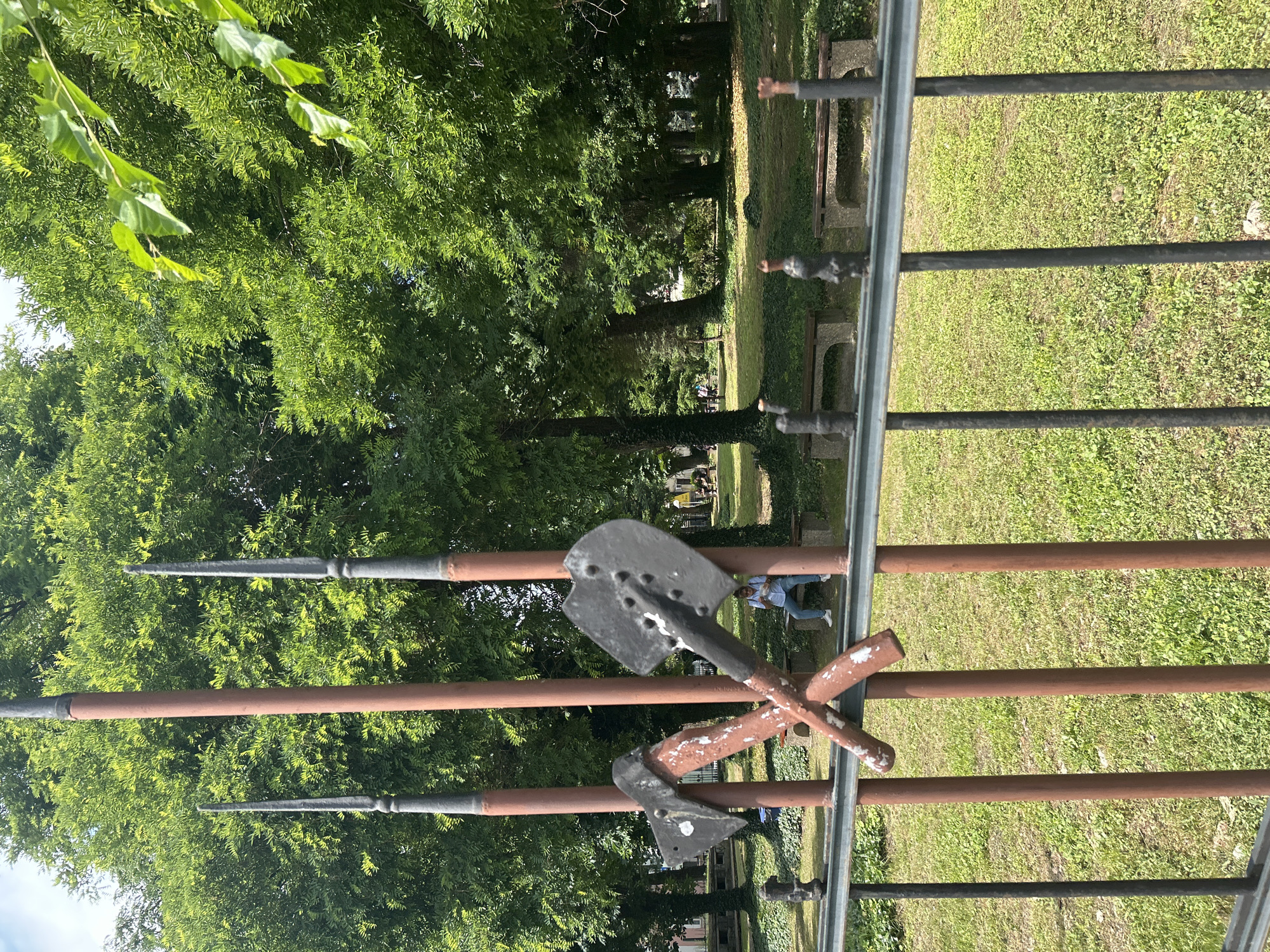 The image displays an outdoor scene, likely a park or memorial area in Pleven, Bulgaria, viewed through an ornate metal fence. The foreground is dominated by this fence, which features dark vertical posts and brown or rust-colored horizontal bars. Attached to the fence are decorative, weathered metallic elements: two spear-like finials extend upwards, and in the center, a grey, stylized spade or shovel is crossed with an axe or pickaxe. These tools show signs of age, with chipped paint.
Beyond the fence, a vibrant green landscape unfolds under bright daylight, suggesting a clear and sunny day. The background is filled with lush, dense green foliage from numerous trees, indicating a thriving natural environment in spring or summer. The ground is covered with neatly kept green grass. Several park benches, appearing to be made of stone or concrete, are scattered across the grassy area in the middle distance.
A person is seated on one of these benches towards the right side of the frame, dressed in blue jeans and a light-colored top, enjoying the peaceful surroundings. Their features are not clearly discernible due to distance. Another faint figure is visible further back among the trees. The scene conveys a sense of calm and leisure in a well-maintained public space, with the decorative fence elements potentially hinting at historical or symbolic significance relevant to the location. No text is visible in the image.
