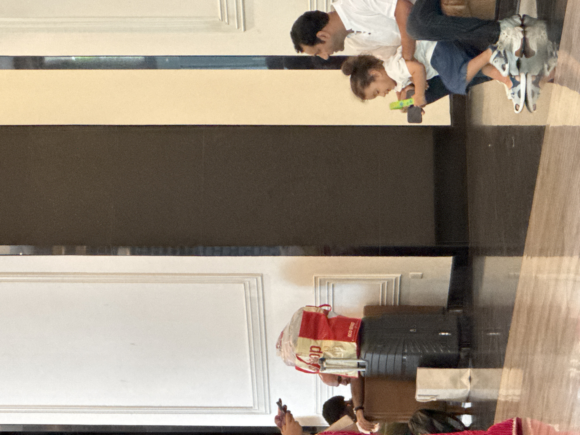 The image depicts a father and his young child in what appears to be an indoor setting, possibly a hotel lobby or airport terminal in Kuala Lumpur, Malaysia. The father, wearing a white t-shirt and dark pants, is holding the child, who is dressed in a white t-shirt and blue shorts. The child is looking down at a toy in their hand. In the background, there is a dark, reflective surface, and further back, a white wall with decorative molding. To the right, a black suitcase and a red and white shopping bag are visible. The overall lighting suggests it could be daytime. The scene appears calm and mundane, with no strong emotional cues visible. The text visible on the shopping bag reads "eco drop" and possibly "shop".