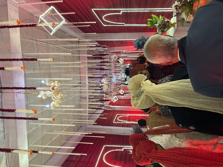 A wedding reception showcases decorative elements. Overhead, red-trimmed white fabric spans the ceiling, adorned with hanging strands of white and maroon floral garlands interspersed with illuminated cube lights and chandeliers. Attendees, some wearing traditional turbans in shades of red, gold, and blue, are partially visible in the foreground, contributing to the celebratory ambiance.