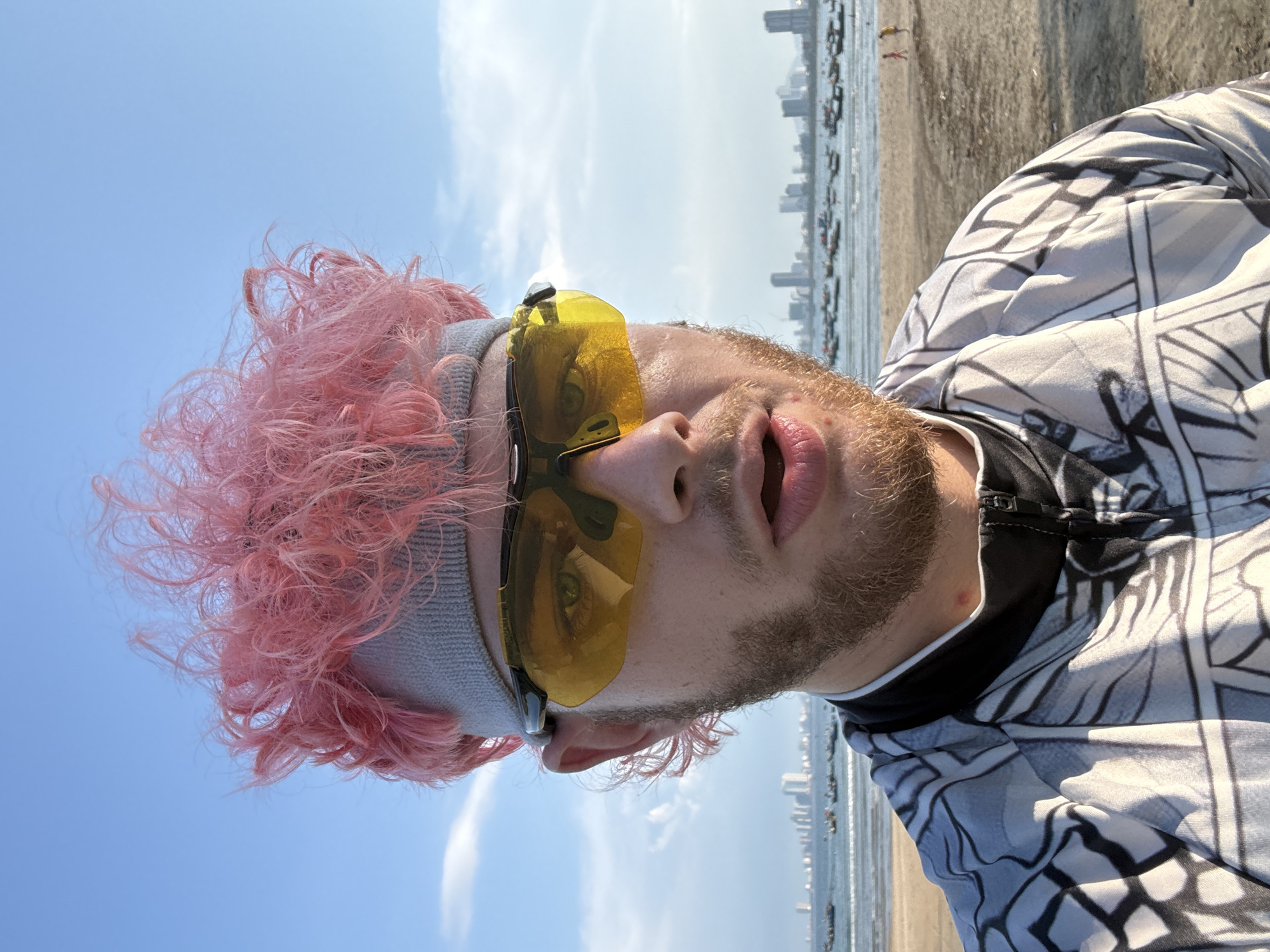 A photograph shows a man with pink hair and a grey headband standing on a beach in Đà Nẵng, Vietnam. He is wearing yellow sunglasses and a patterned jacket. The background shows the beach, the sea, and the skyline of Đà Nẵng.