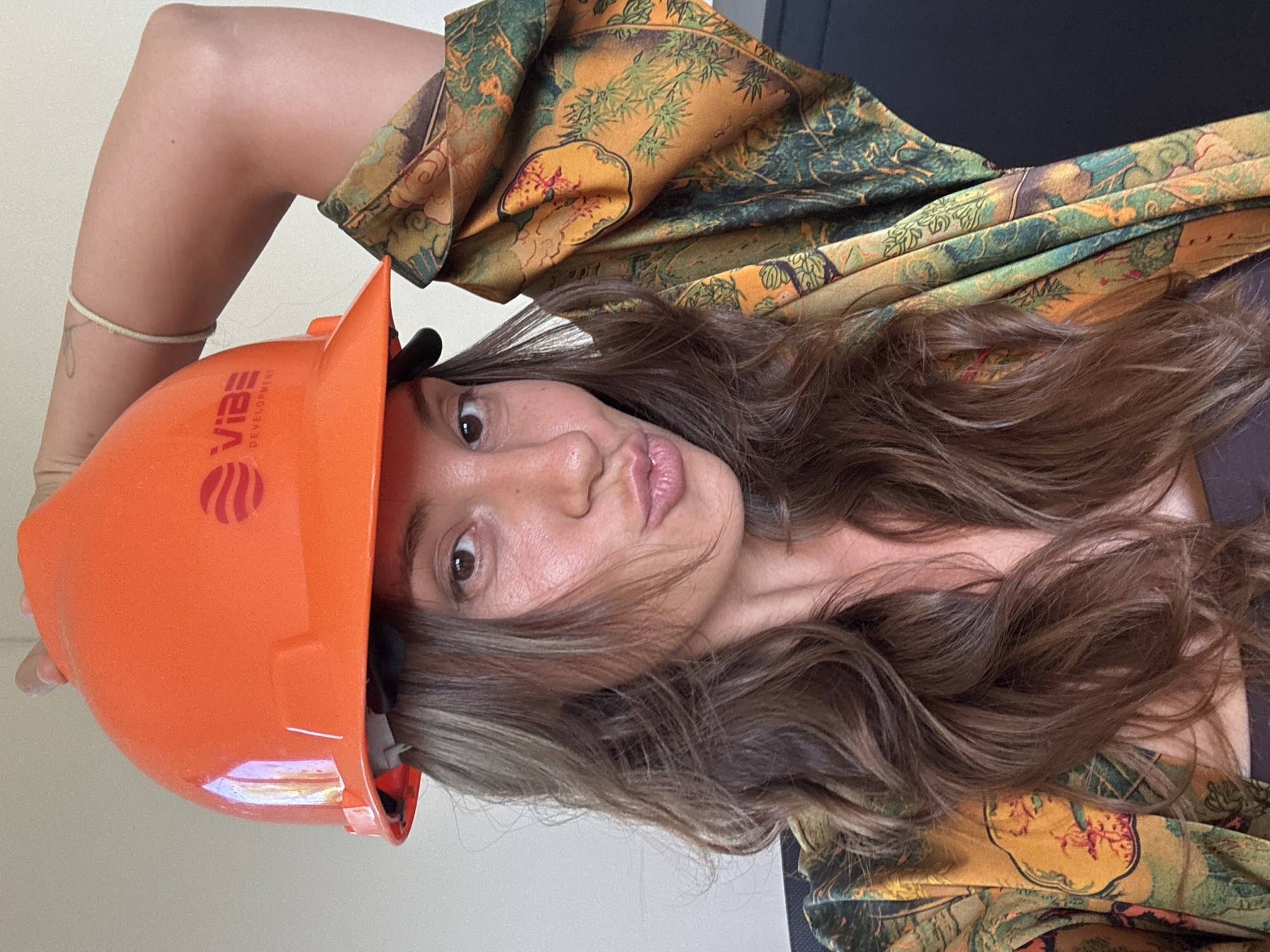 A woman with light skin, long brown hair, and brown eyes is holding an orange hard hat with the word "Vibe Development" on it above her head, tilted to the side. She is wearing a patterned green and yellow shirt. The background is plain white. The location is not discernible as Kuta Selatan, Indonesia based on the image provided. There are no other people or significant objects visible. The woman is making a kissing face.