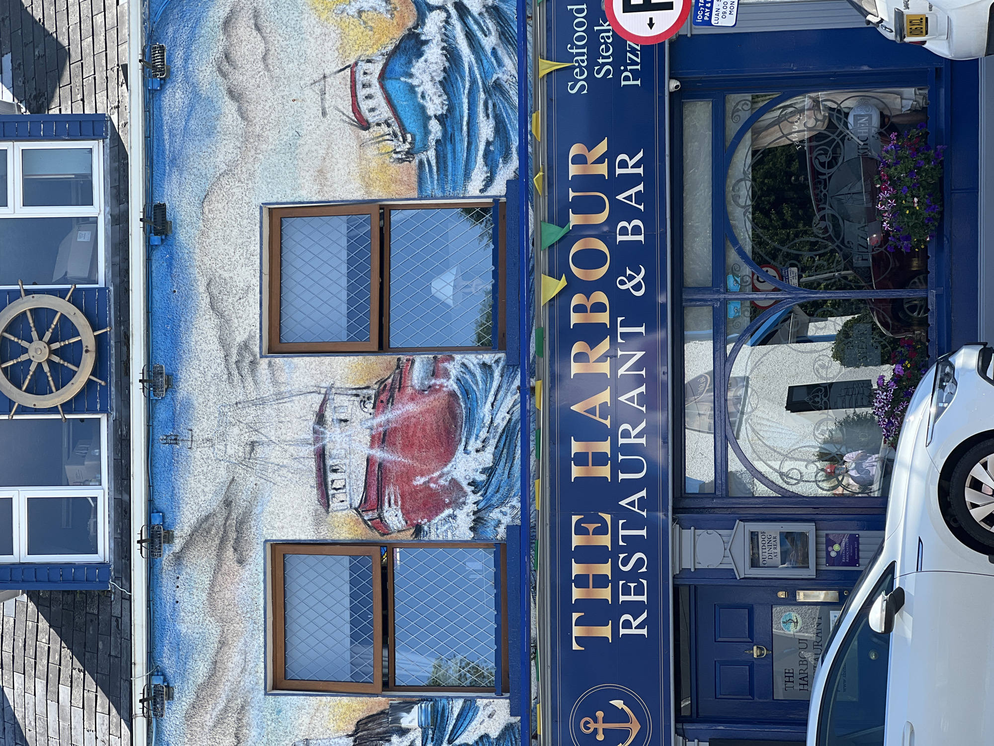 The image depicts the facade of "The Harbour Restaurant & Bar" in Donegal Town, Ireland. The building's exterior is adorned with a large mural of ships sailing on a rough sea under a cloudy sky. Two upper-story windows with diamond-patterned glass are visible within the mural. Below the mural, a dark blue awning displays the restaurant's name in gold lettering, accompanied by an anchor symbol on the left. The awning also lists menu items such as "Seafood," "Steak," and "Pizza."
The ground floor features a blue door on the left, with a sign indicating "The Harbour Restaurant" and a small anchor logo. To the right of the door is a display case for "Outdoor Dining at Rear." A large arched window with decorative etching and a security camera is to the right of the entrance. Inside the window, a red car and plants are visible. A white car is parked in front of the building, partially obscuring the lower portion of the facade. A traffic sign with a red circle and a white 'P' is attached to a blue pole to the right of the restaurant entrance. The architectural style suggests a traditional building, likely located in a coastal town given the nautical theme.