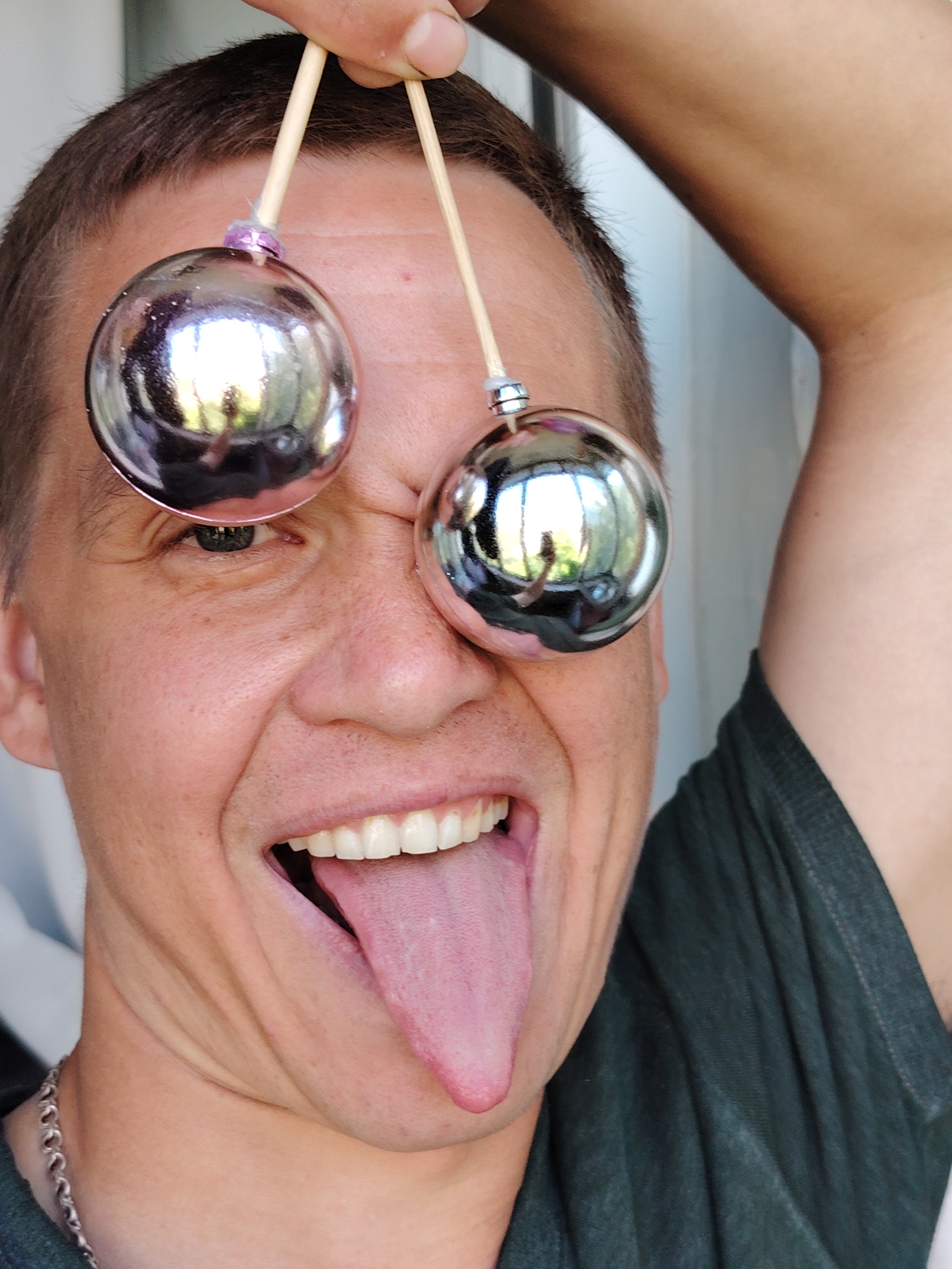 In a close-up, vertical shot, a person's face is the central focus, with their head tilted to the side and their mouth wide open, showing their teeth and a long, pink tongue sticking out. Two shiny, spherical ornaments, likely Christmas tree decorations, are held up on wooden skewers, positioned just above the person's eyes, obscuring them from view and reflecting the surrounding environment. The person is wearing a dark green t-shirt and a silver chain necklace. The background is blurred, suggesting an indoor setting with diffused light, possibly near a window, as evidenced by reflections in the ornaments. No specific location within Chaikovsky, Russia, can be determined from the image itself.
