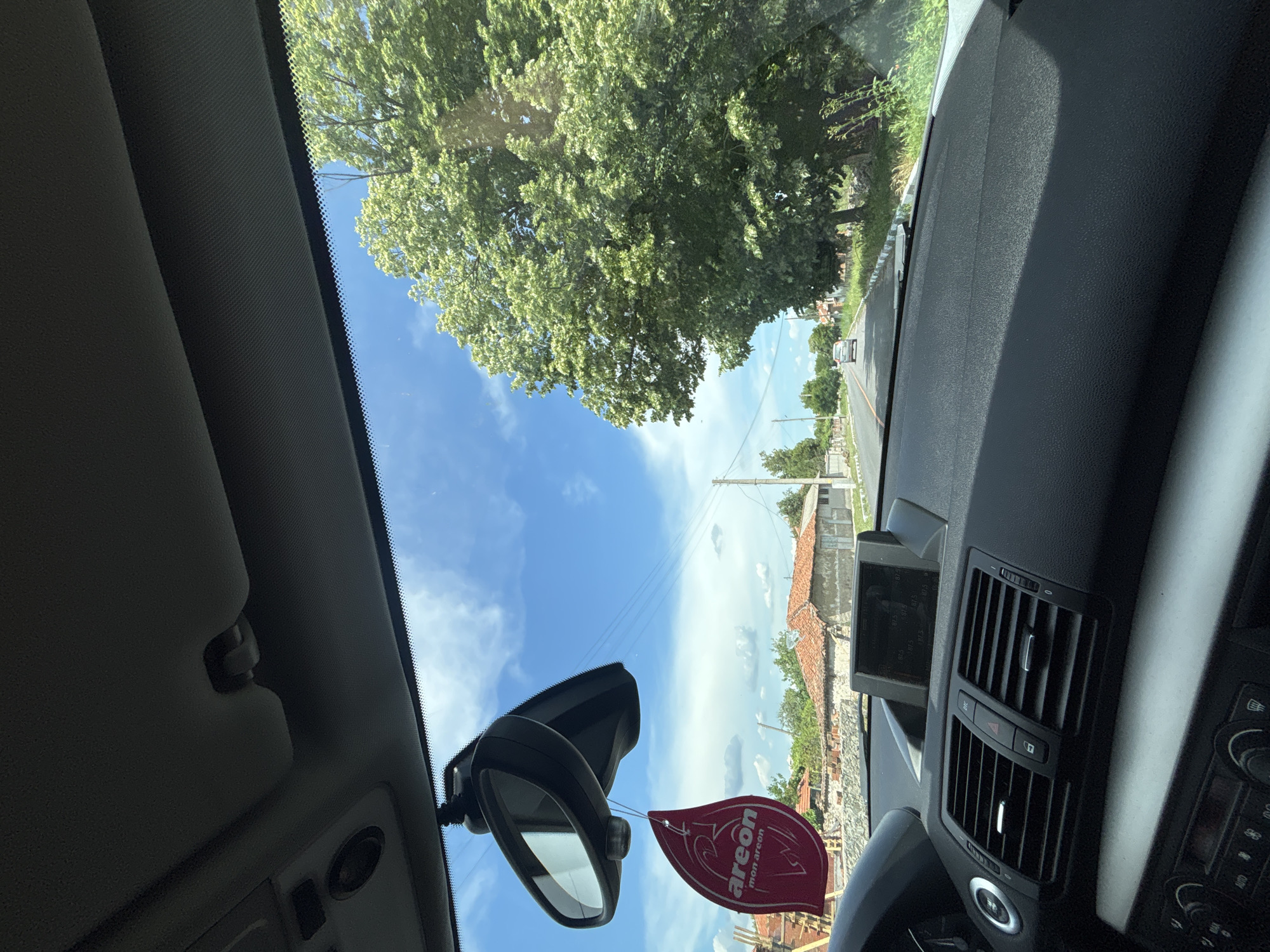 The image is a sideways, wide-angle shot taken from inside a car, looking out through the windshield. The scene depicts a bright, sunny day with a clear blue sky dotted with wispy white clouds. Lush green trees, their leaves illuminated by sunlight, dominate the upper portion of the frame. A road is visible, with a car in the distance and trees lining its sides. Houses with red tile roofs and some construction are also present, suggesting a rural or semi-rural setting.
Prominently hanging from the rearview mirror is a red, leaf-shaped air freshener with white text. The text reads "areon" and "mon areon". The interior of the car is visible in the foreground, including parts of the dashboard, air vents, a sun visor, and the steering wheel. The overall impression is of a pleasant drive on a summer day. The location cues, such as the architecture of the houses and the style of the road, along with the provided location context, suggest the setting is Varbitsa, Bulgaria. The lighting indicates it is daytime, likely late morning or early afternoon.