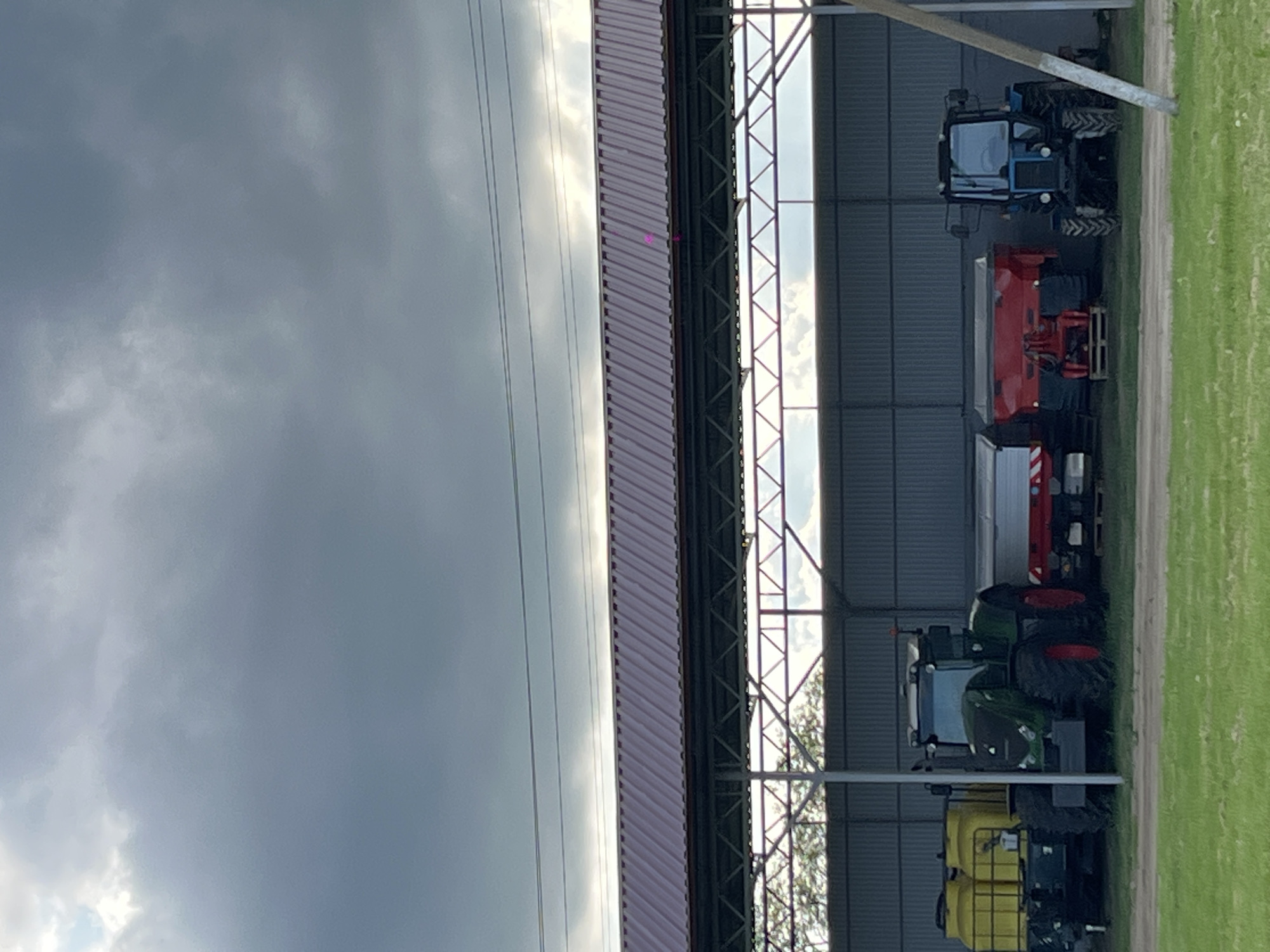 The image shows a sideways view of a building, likely a storage shed, in Novopokrovskaya, Russia. Inside the shed, several pieces of farm equipment, including tractors and other machinery, are visible. Outside, there is a grassy area. The sky is overcast, suggesting potential rain. No people are visible.