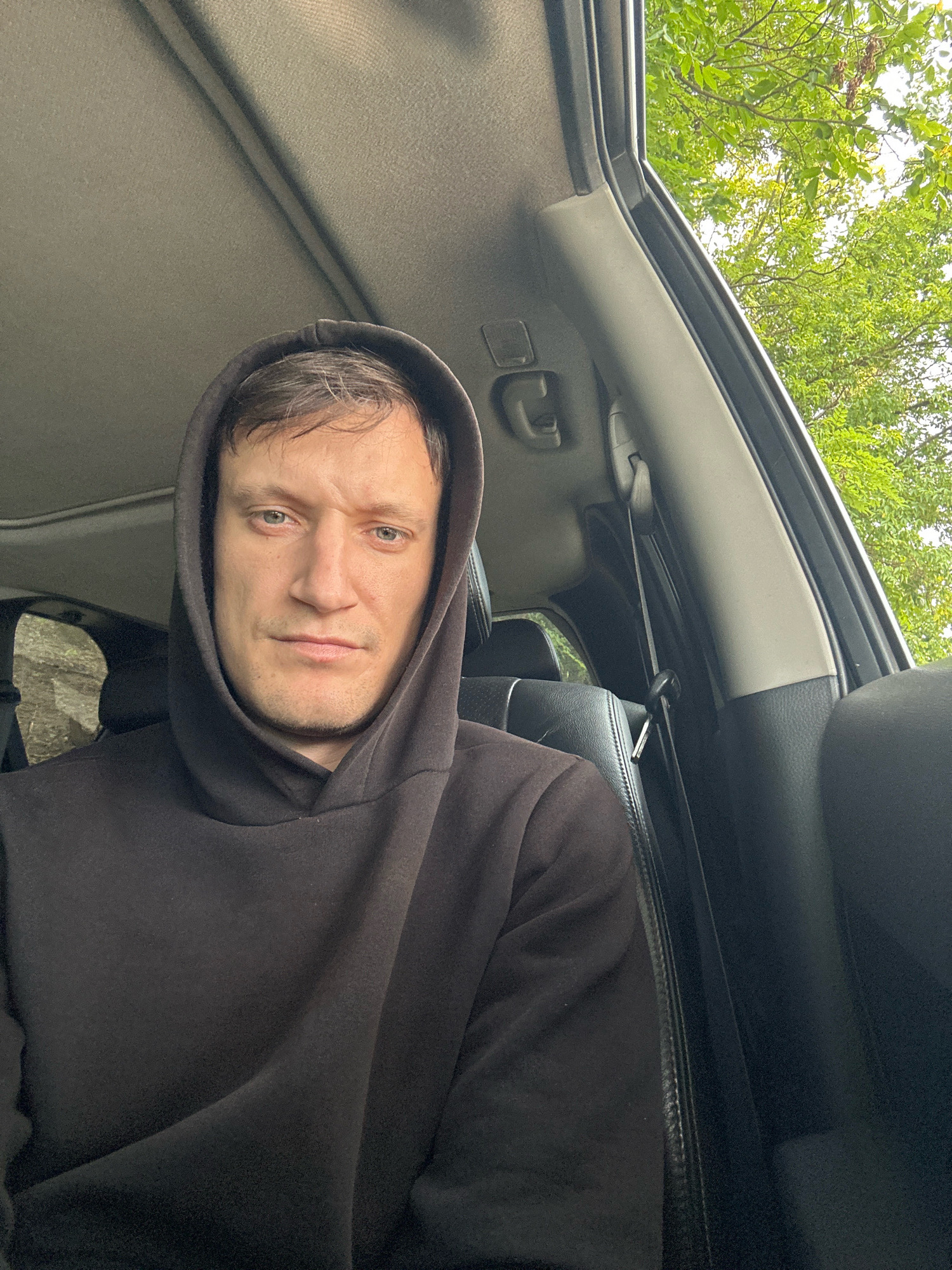 A close-up, selfie-style shot shows a man wearing a dark hoodie while sitting in the passenger seat of a car. The man's face is centered in the frame, and he is looking directly at the camera with a neutral expression. His hood is up, obscuring the top of his head. The interior of the car is visible, including the headliner and a portion of the dashboard. Through the car window, green foliage and branches of trees are visible, suggesting an outdoor setting, possibly on a street or in a park. The lighting appears to be natural daylight. There is no indication of the man's location within the city of Makiv, Ukraine, or any other specific geographical context.