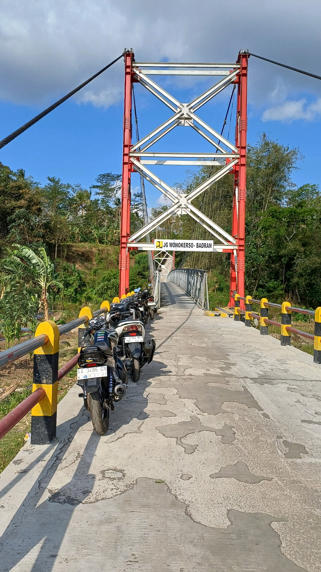 The image shows a suspension bridge in Wonokerso, Indonesia, with several motorcycles parked on its pathway. The bridge features a prominent red and white steel structure supporting the suspension cables. The pathway is made of concrete and is lined with yellow and black striped safety barriers. Lush green trees and vegetation surround the bridge. A sign on the bridge reads "JG WONOKERSO - BADRAN". The sky is blue with some scattered clouds, indicating a clear day. The perspective is from ground level, looking across the bridge.