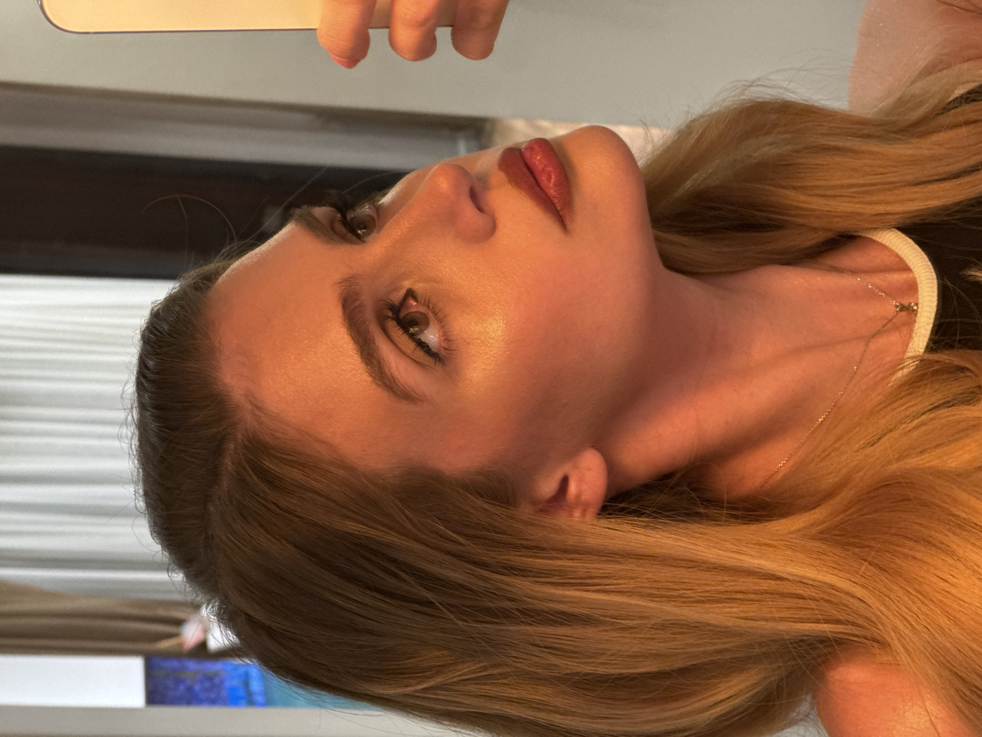 The image captures a close-up, rotated portrait of a woman, featuring her face and upper body from a high angle. Her head is tilted, giving the impression she is looking upwards and to the right. She has long, flowing blonde hair, with visible darker roots at the part, cascading around her shoulders and neck.
Her fair skin is well-lit, highlighting her features. She has defined eyebrows and visible eyelashes framing her brown eyes, which gaze upwards. Her lips are painted a rich reddish-brown shade, appearing slightly parted or in a gentle pout. A delicate, thin chain necklace is visible around her neck, resting on her skin and partially covered by her hair. A white, ribbed strap of what appears to be a top or undergarment is visible on her right shoulder.
At the top of the frame, a finger and a section of a light-colored, reflective object (possibly a phone or mirror) are visible, suggesting the image may be a selfie or taken in front of a mirror. The background is softly blurred, showing horizontal lines, possibly from blinds or slats, on the left, and darker, indistinct shapes elsewhere, indicating an indoor setting. The lighting is strong and directed, creating highlights on her cheekbones and forehead, giving her skin a radiant appearance. There is no visible text in the image.
