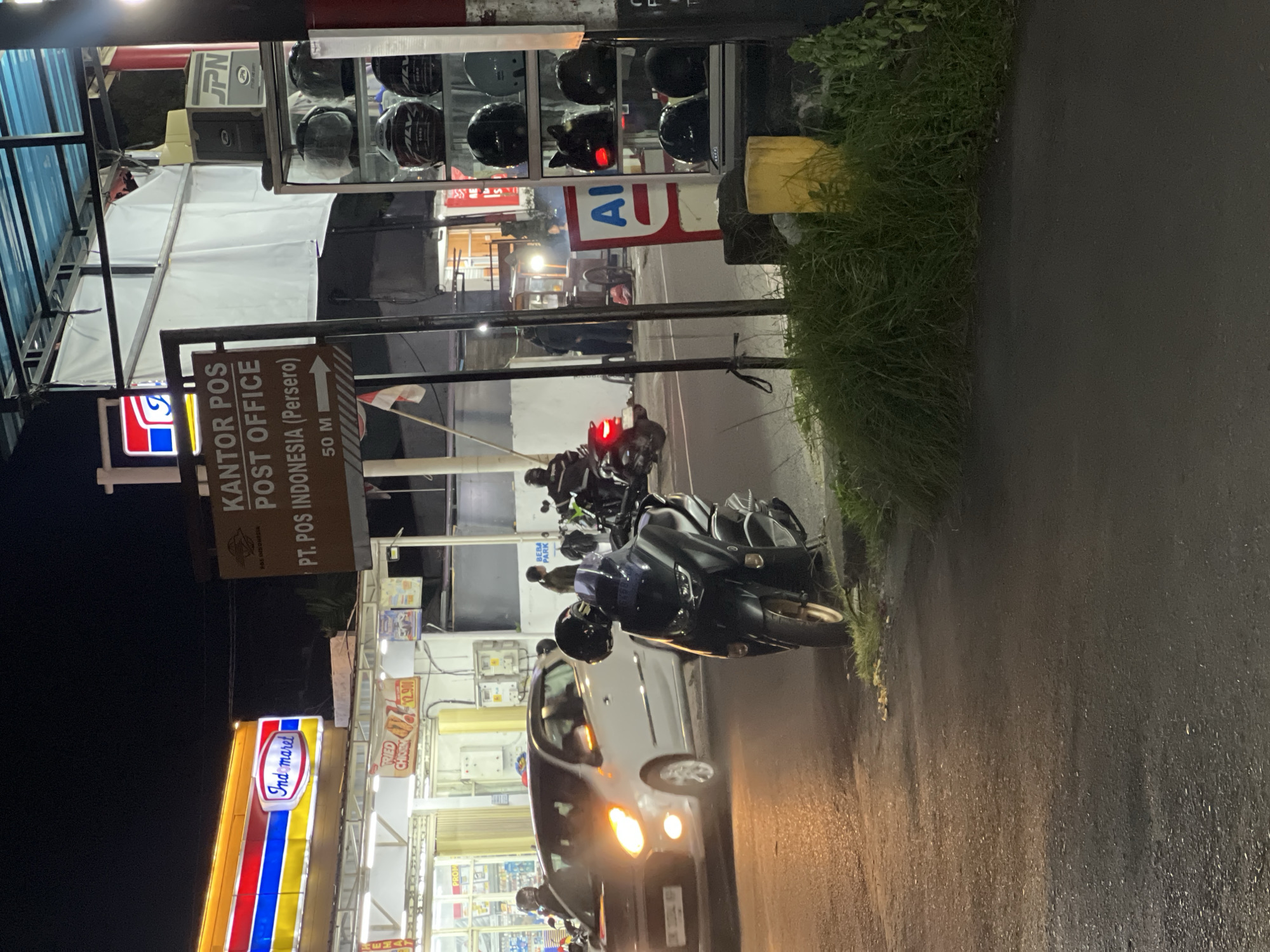 This nighttime street scene, likely in Gianyar, Indonesia, captures a bustling corner outside an Indomaret convenience store and a PT. POS Indonesia (Persero) post office. A white car with its headlights on is stopped on the asphalt road, with a dark-colored motorcycle parked on the grassy verge in the foreground. The post office sign, with "KANTOR POS POST OFFICE PT. POS INDONESIA (Persero) 50 M" clearly visible, points towards the left. Behind the motorcycle, shelves display several helmets. In the background, the brightly lit Indomaret sign is prominent, with various items displayed inside the store. The environment is characterized by artificial lighting illuminating the street and buildings, with dark skies above. Several individuals are visible in the background, some near the Indomaret and others near motorcycles. The overall impression is of a lively urban environment after dark, with commercial activity and transportation present.