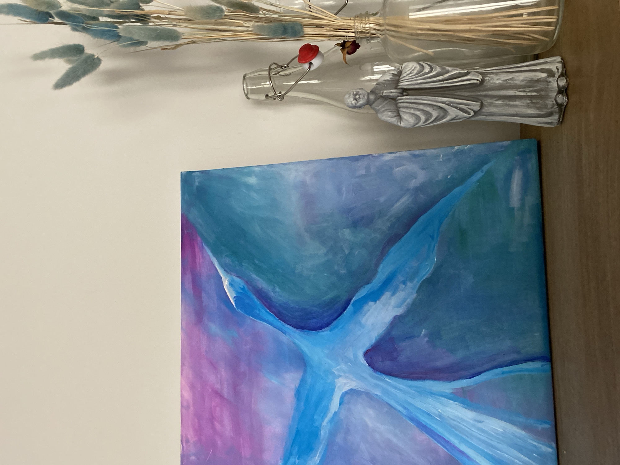 A painting, predominantly blue and purple with a white, angular design in the center, rests against a white wall and a light brown, wooden surface. Above the painting, a glass bottle lies horizontally, partially filled with dried grasses. Another smaller glass bottle, sealed with a metal clasp and a red top, contains a small figure made of a grey material. Some dried blue foliage is located near the bottles. The city of Lâm Viên - Đà Lạt, Vietnam, is not depicted or suggested in the scene.