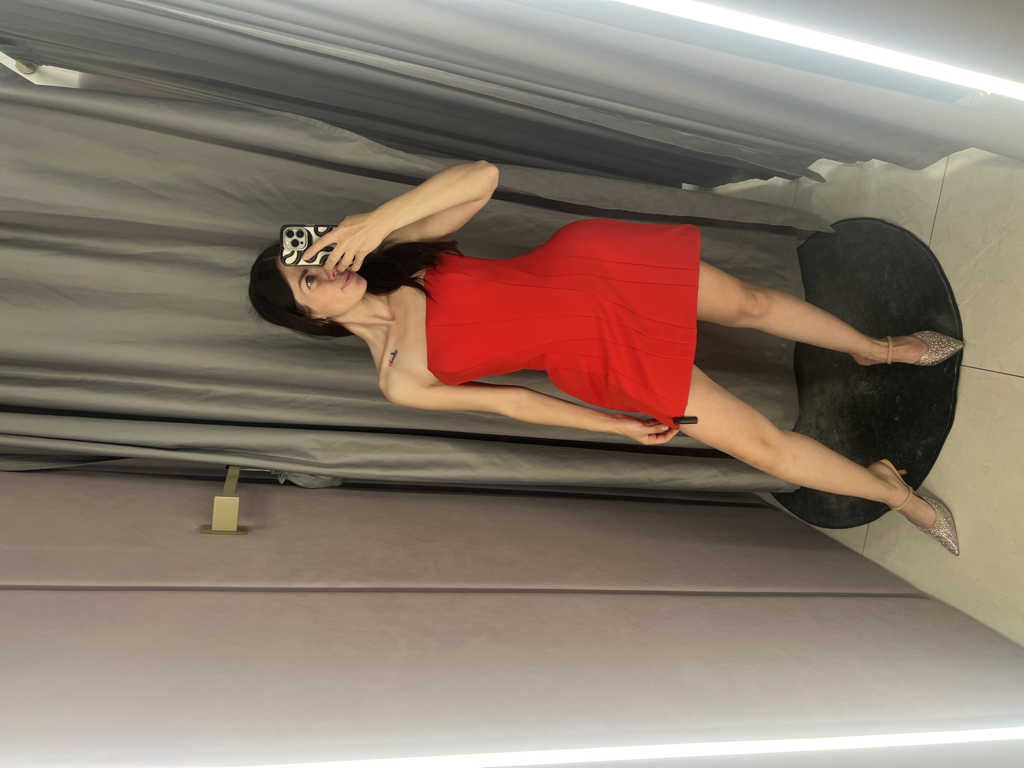 A woman is standing in a dimly lit fitting room, taking a selfie in a mirror. She is wearing a strapless red dress and sparkly silver heels. The fitting room has grey curtains and light-colored walls, with bright vertical lights on either side of the mirror. A gold hook is attached to the wall on the left. The floor is tiled and there is a black circular rug under the woman's feet. The woman's hair is dark brown and she has a tattoo on her left shoulder. The image appears to be taken in a retail setting, possibly a clothing store.