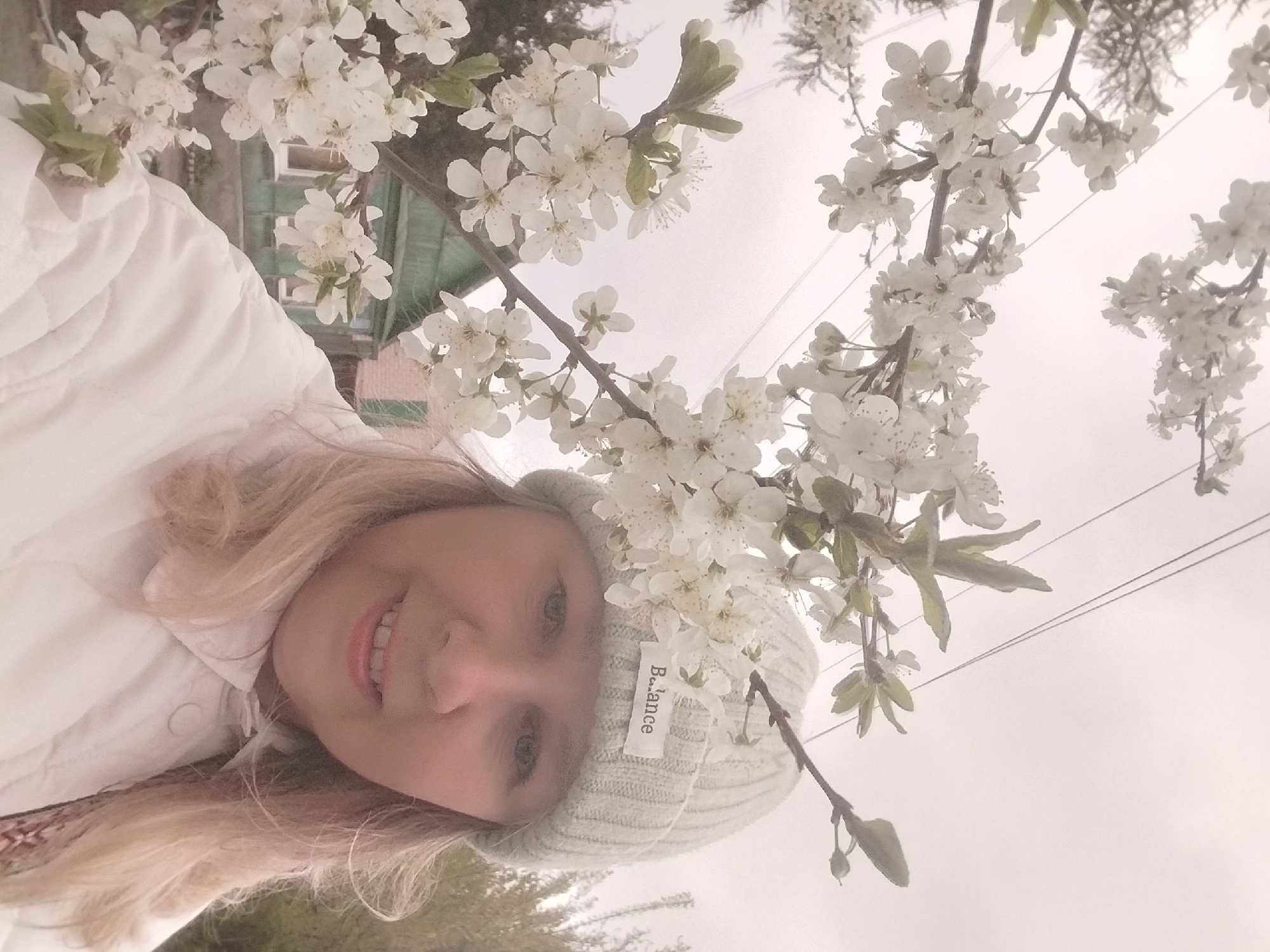 A blonde woman with a bright smile looks towards the camera, captured in a close-up, slightly tilted portrait. She wears a white quilted jacket and a light grey, ribbed beanie that has a white tag with "B..lance" written on it. Her eyes are light-colored.
The woman is surrounded by numerous branches of a blossoming tree, likely a cherry or plum, with an abundance of small white flowers framing her head and the upper part of her body. The full bloom indicates that the photo was taken in spring. The background consists of a bright, overcast sky, giving the scene a soft, diffused light. Parts of a green-colored building are visible in the upper left background, suggesting a residential or garden setting. Thin power lines can be seen extending across the top right portion of the frame. The overall mood is cheerful, reflecting the woman's smile and the vibrant spring blossoms. This scene is set in Orenburg, Russia.