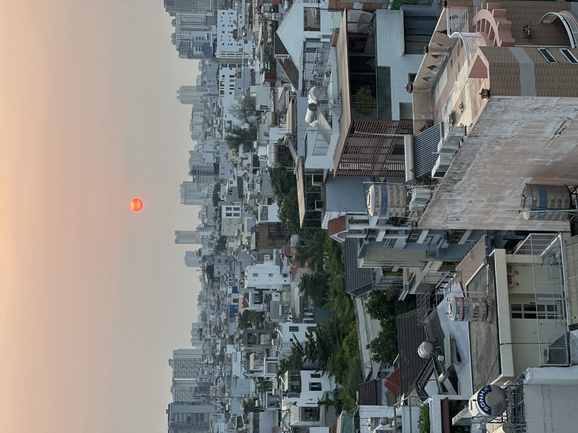 The image depicts a dense urban landscape in Ho Chi Minh City, Vietnam. The view is captured from an elevated perspective, overlooking a mix of low-rise houses and taller apartment buildings. The structures are tightly packed, creating a visually complex scene with varying architectural styles. In the distance, a hazy sky is visible with the sun.