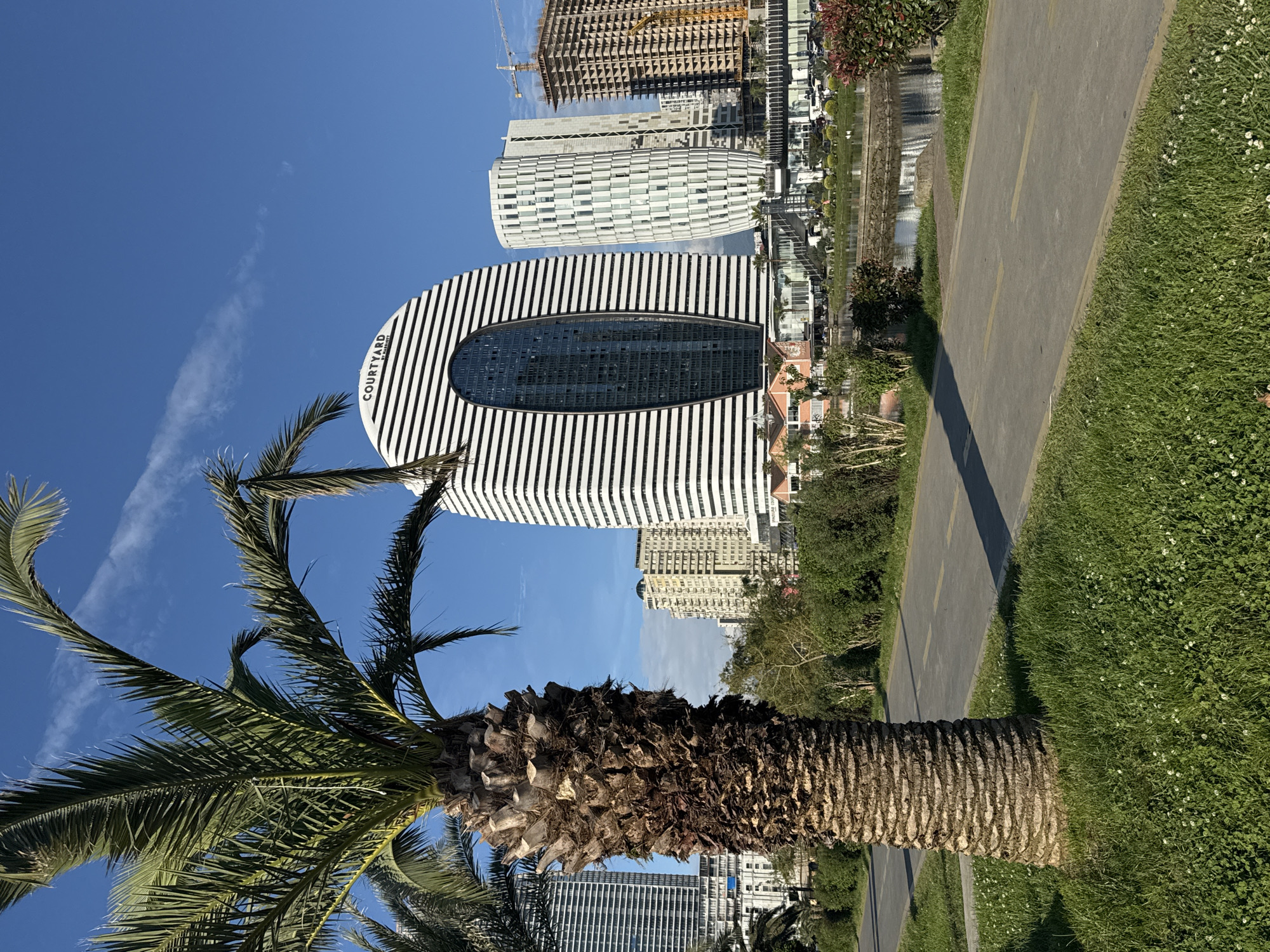 The image shows a view in Batumi, Georgia. A palm tree is in the foreground on a grassy lawn. Behind the tree is the Courtyard by Marriott hotel, a distinctive oval-shaped building with black and white horizontal stripes and an oval window. Other buildings, including residential towers, are visible in the background, along with a construction crane. A paved pathway runs along the edge of the lawn. The sky is blue with some wispy clouds.