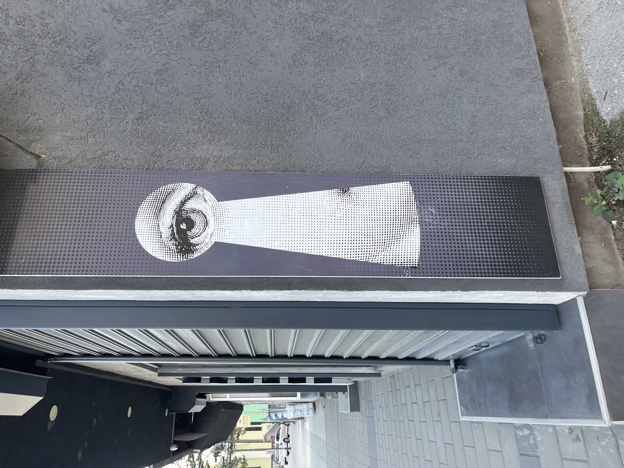 This image captures a section of a building's facade on a sidewalk in Cacak, Serbia. The most prominent feature is a vertical poster on the right side of the image, displaying a black and white graphic resembling a keyhole. Within the top, circular part of the keyhole, a detailed human eye is depicted, with the iris rendered through intricate line work and shading. The lower, elongated part of the keyhole is filled with a gradient of black dots, transitioning from solid black at the bottom to white at the top, where it meets the eye.
To the left of the poster, a gray metal roll-up security shutter is visible, partially obscuring the entrance to a building. The shutter is in a closed position. Above the shutter, a dark, angled overhang of the building is present, with recessed lighting fixtures. The sidewalk in front of the building is paved with gray square tiles. The overall environment appears to be an urban setting, likely a commercial area. The wall on which the poster is affixed has a rough, textured surface. There are no people or vehicles directly in the frame of this specific image, but a street and parked cars are visible in the background to the far left.