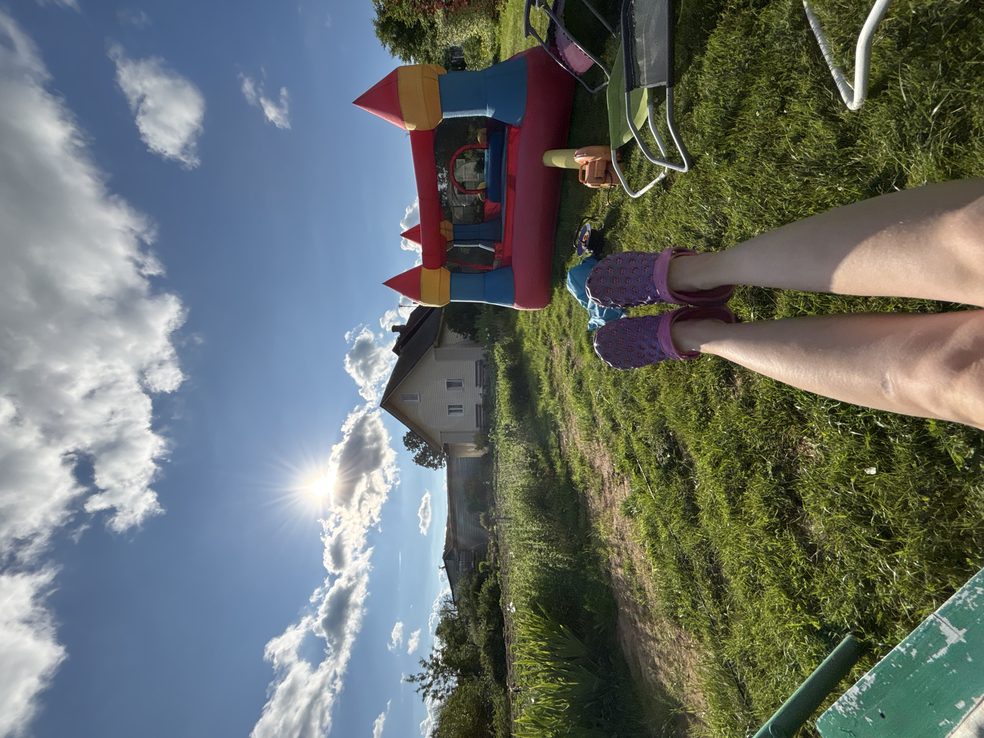 The image depicts a sunny outdoor scene in the village of Dunilovo, Russia. In the foreground, a person's legs, wearing purple patterned clogs, are extended towards the viewer. The ground is covered in lush green grass. To the right, a colorful inflatable bounce house is set up. Various items are scattered on the grass around the bounce house, including what appears to be a leaf blower, a blue bag, and parts of lawn chairs. In the background, a light-colored house with a prominent roofline stands. Beyond the house, there are trees and other vegetation, suggesting a rural or semi-rural setting. The sky is blue with scattered white clouds, and the sun is shining brightly, casting rays of light.