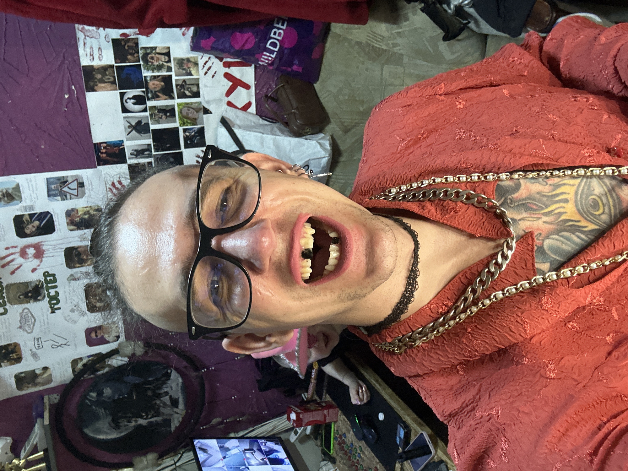 The scene takes place indoors, stated to be within the city of Saratov, Russia; however, no specific geographic identifiers are visible in the image itself. The primary subject, an adult individual, is centered in a selfie, looking directly at the viewer with a wide-open mouth revealing missing upper and lower central incisors, and discolored or decaying teeth, notably a dark tooth on the lower left. The individual wears black-rimmed glasses, a red textured garment (possibly a robe or shirt), a black lace choker, and multiple gold-colored chains. A prominent tattoo depicting a flaming eye within a shield or heart shape is visible on the chest. The subject's left ear displays a silver hoop earring and a silver, key-shaped earring.
In the background to the left, a second individual, appearing to be a child, is partially visible, wearing a pink hat or head covering and leaning over a desk. On this desk, a monitor displays a grid of multiple video feeds. Other objects on the desk include a red box, a mobile phone, a computer mouse, a small white cylindrical object, and numerous small, colored, round items scattered on a dark mat.
The wall behind the subjects is heavily adorned with a collage of photographs and papers, including a large, dark-framed circular picture featuring two faces, a red handprint, and various images with Russian text, such as "ГОСТЕВ" and "ЛАС ВЕГАС," along with a road sign. Sections of the wall appear purple or burgundy. To the right, multiple bags are visible on the floor or a surface, including a purple bag with "WILDBE" branding (likely Wildberries). A red fabric, consistent with the primary subject's garment, is draped on the far right. The interior space is brightly lit, consistent with artificial lighting.