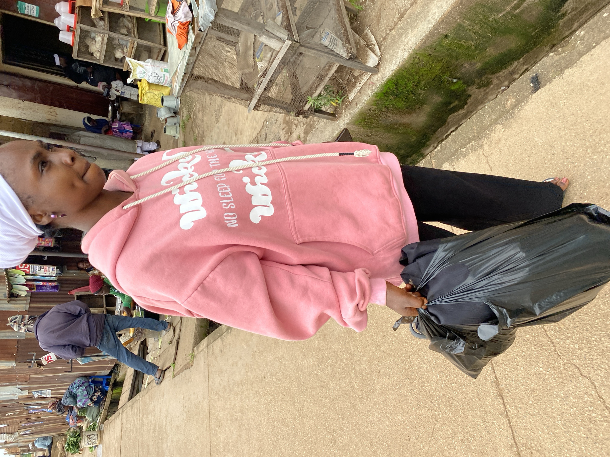 In Bukuru, Nigeria, a young Black woman wearing a pink hoodie and black pants stands on a concrete walkway, holding a large black plastic bag filled with refuse in her right hand. The background reveals a bustling marketplace. To her left, a man in a blue jacket and jeans walks away, carrying produce. Further back, women sit with market wares. To her right, a weathered wooden structure, possibly a market stall, displays shelves containing various items, including bags of grain and poultry feeders. The environment appears somewhat unkempt, with visible moss growth along a concrete drainage channel to the right. The overall scene captures a moment of daily life in a Nigerian urban market setting.