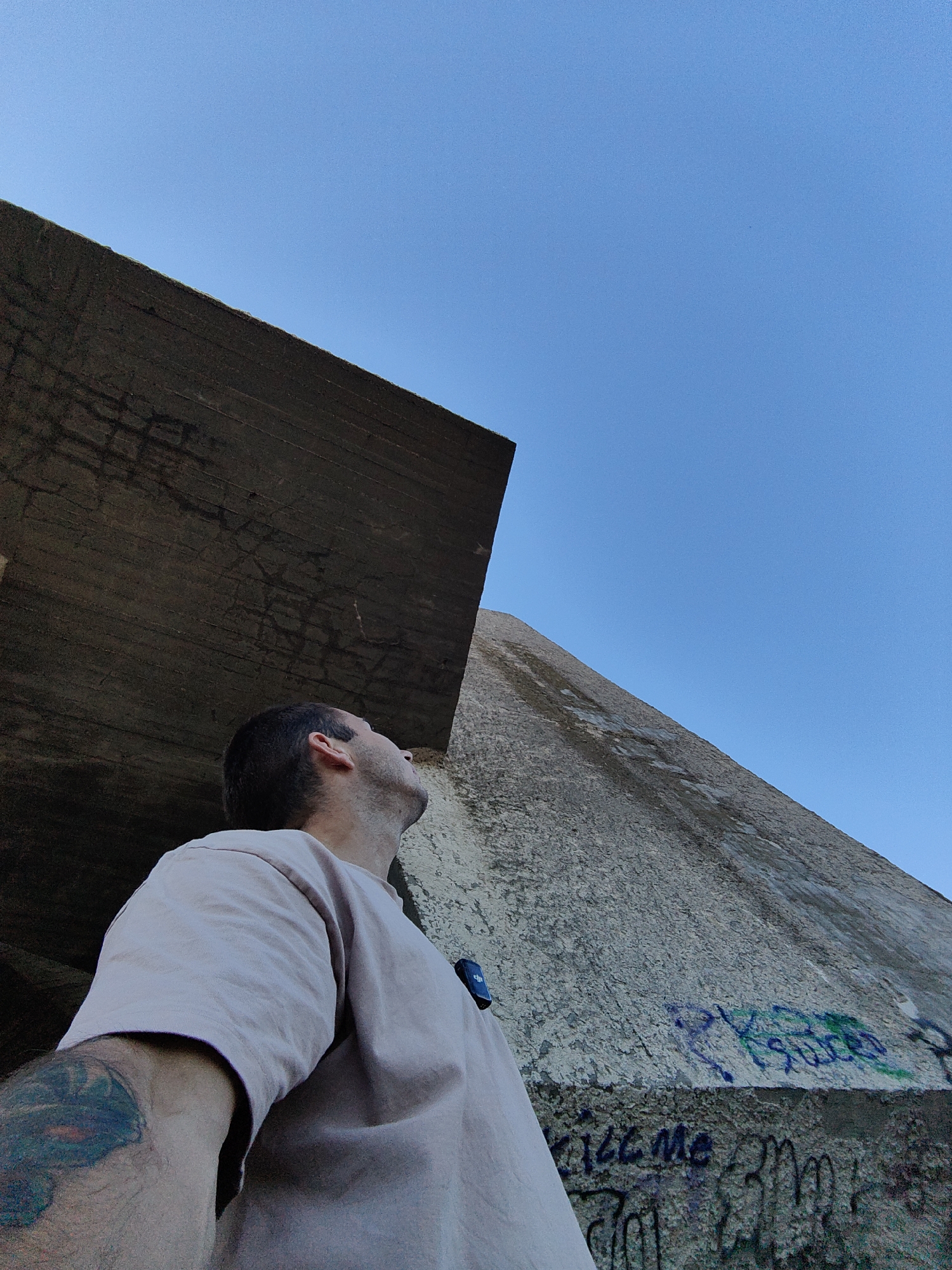 A low-angle, full shot captures a man from the chest up, looking upwards, situated in front of imposing, weathered concrete structures. The man, wearing a light-colored t-shirt, has a visible tattoo on his left arm. To his left, a dark, textured concrete surface juts out diagonally from the upper left corner towards the center, with visible lines and cracks. To his right, a lighter, rougher concrete wall slopes upwards into the bright, clear blue sky. This wall has graffiti scrawled across its lower section. The scene appears to be outdoors, with the stark concrete contrasted against the expansive sky. It is plausible that this image was taken in the vicinity of the Varna Shipyard or other industrial areas along the Varna Bay, known for their large concrete structures and potential for graffiti.