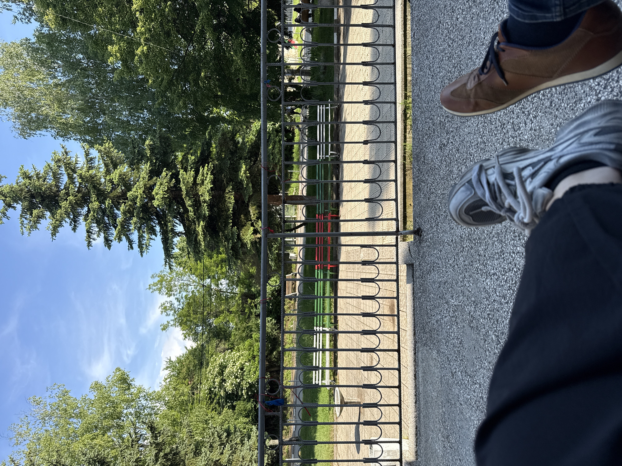 The image captures an outdoor scene in Dragichevo, Bulgaria, from a low-angle, first-person perspective looking into a public park on a clear day.
In the immediate foreground, the lower legs and feet of two individuals are visible. On the left, a leg clad in dark fabric wears a grey athletic shoe with a textured, possibly ridged, upper and matching grey laces. On the right, a leg covered by blue denim fabric wears a brown leather or suede lace-up shoe with dark laces and a light sole, exposing dark socks. The ground directly beneath the feet is a light grey, speckled aggregate surface.
A dark grey, wrought-iron fence extends horizontally across the mid-ground, acting as a partition. The fence features decorative elements including horizontal bars, vertical spindles with U-shaped bases, and circular motifs along the top rail. Several small red and blue ties or ribbons are affixed to the top of the fence.
Behind the fence, a public space unfolds, featuring a paved area made of light-colored square or rectangular tiles. Three park benches are aligned in a row on this paved section: a white bench on the left, a green bench in the center, and a red bench on the right. To the left of the benches, a stone structure resembling a birdbath, with a wide, shallow basin on a pedestal, is visible.
The background is dominated by lush green trees under a bright blue sky with scattered wispy white clouds. Tall, dark green conifer trees are prominent on both the left and right sides of the frame, flanking various deciduous trees with lighter green foliage in the central background. Some deciduous trees exhibit visible white bark. The sunlight is strong, illuminating the scene and creating shadows. In the deeper background, indistinct human figures can be observed walking or sitting on other benches within the park area.