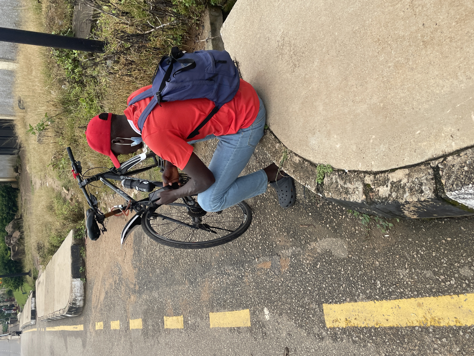 A person is sitting on a concrete curb next to a bicycle on a street in Jos, Nigeria. The person is wearing a red t-shirt, blue jeans, a red baseball cap, and a blue backpack. A light blue face mask hangs from the person's ear. The bicycle is black with a water bottle attached to the frame. The street has yellow lane markings. In the background, there is dry grass, bushes, rocks, and a building. The overall impression is of a person taking a break during a bicycle ride in an urban or semi-urban environment in Jos, Nigeria.