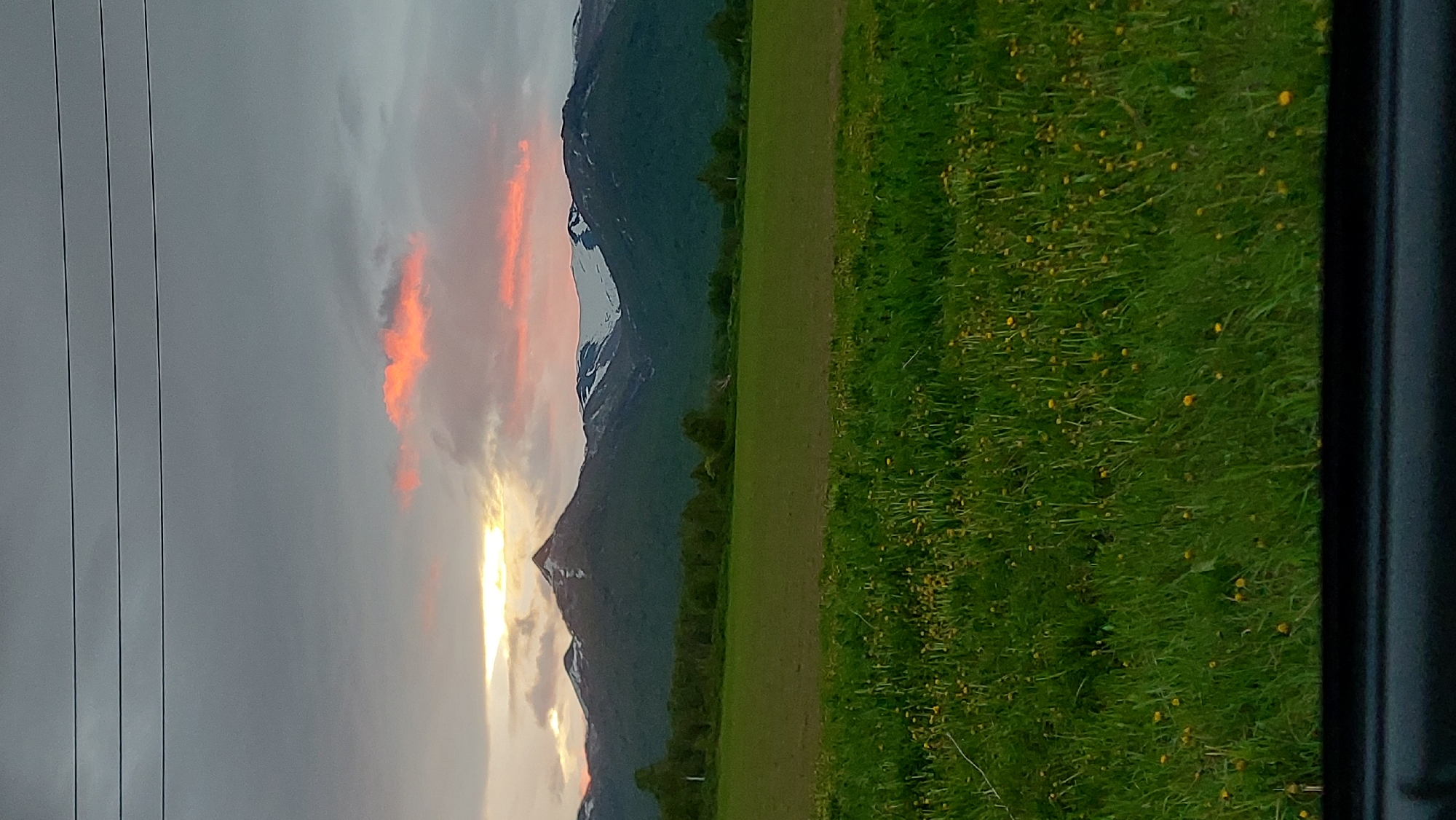 The image depicts a mountainous landscape with a field in the foreground under a cloudy sky. The mountains in the background are partially covered in snow, and the sky is illuminated by an orange sunset. A line of trees separates the field from the mountains. The field itself is lush green with scattered yellow wildflowers. The bottom of the image shows a dark, blurry area, possibly a car interior or frame, suggesting the photo was taken from a vehicle. The scene is devoid of any people or immediate activity. The location in Lesnoi, Russia is not discernable from the visual content of the image alone.