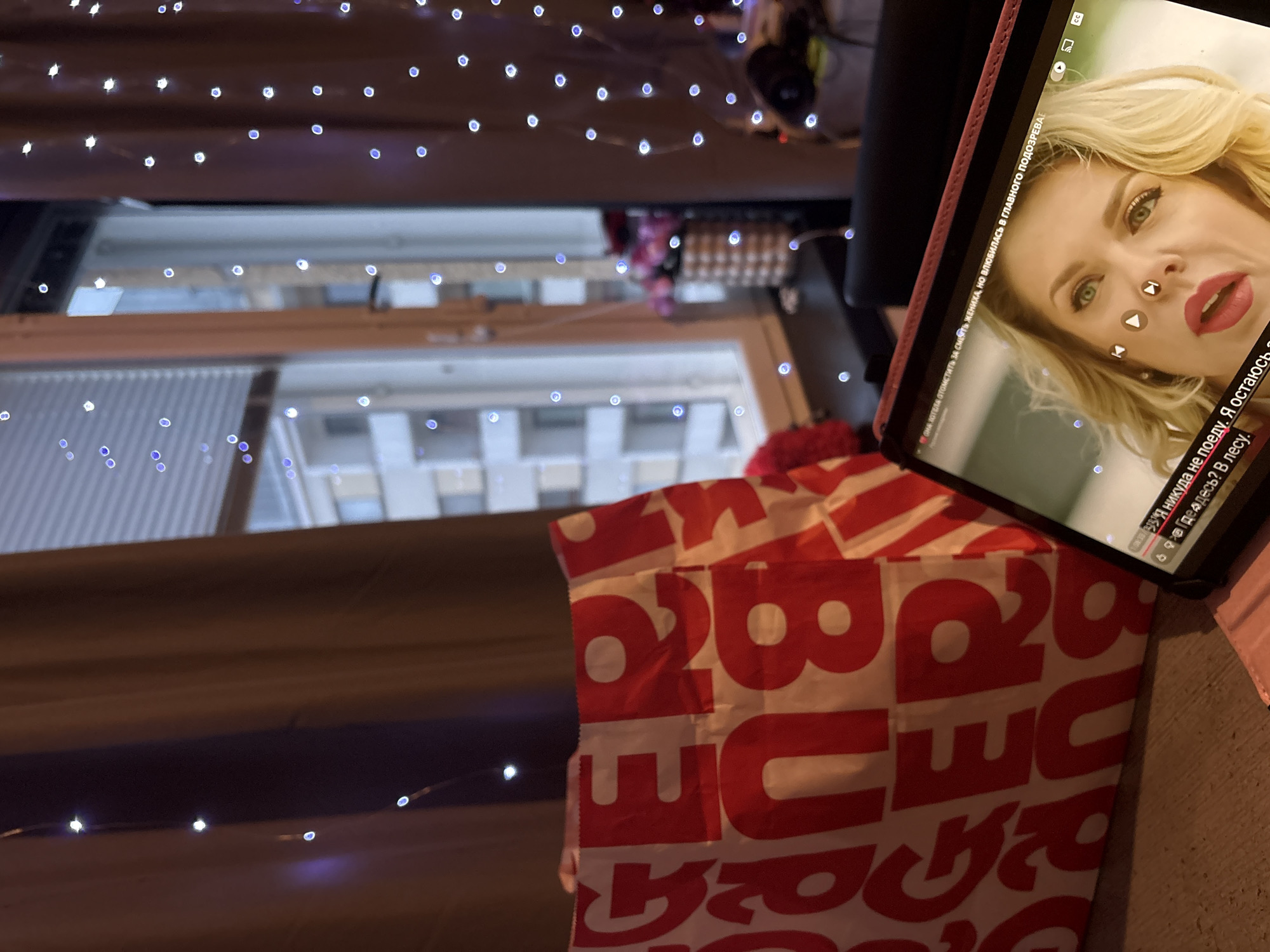 The image shows a cozy indoor scene with a tablet playing a video, a shopping bag with red lettering, and string lights decorating the window area. The tablet displays a close-up of a blonde woman with red lipstick and green eyes. Text on the tablet appears to be in Russian, with phrases like "Я никуда не поеду. Я остаюсь в лесу." visible.
The shopping bag is prominently placed to the left of the tablet and features large red letters, possibly from a fast-food chain. Behind the shopping bag and to the left of the window, a curtain of brown fabric is visible, adorned with a string of small, white, circular lights. The window itself shows a view of buildings and a street, suggesting an urban environment. To the right of the window, more brown curtains and string lights are visible. A small vase with pink flowers is placed on the windowsill. The overall lighting suggests it might be evening or nighttime, with the string lights creating a warm ambiance.
The question of the location being Pori, Finland cannot be confirmed from the visual information provided. The image itself does not contain any specific landmarks or elements that definitively place it in Pori, Finland.