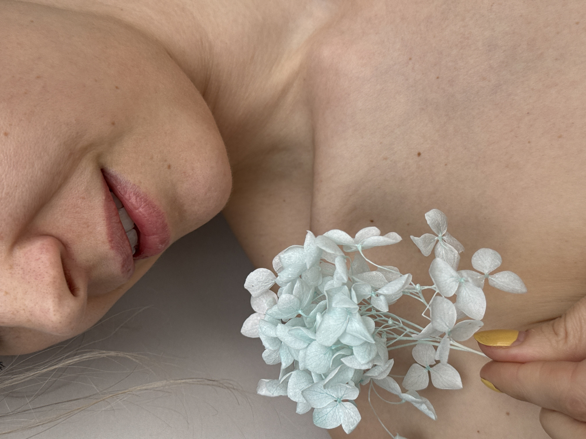 This intimate, high-angle close-up image captures the upper body of a person, likely female, lying down in an indoor setting. The left side of the frame features the person's face, showing her nose, chin, and a slightly parted mouth revealing a hint of teeth, suggesting a soft, contented smile. Her fair skin is visible, adorned with light freckles.
The central and right portions of the image focus on her bare upper torso, including the chest and shoulder area. Held gently against her chest are delicate, light blue flowers, resembling dried hydrangeas, which are the main object in the scene. The fingers of one hand, distinguishable by a short, bright yellow-painted fingernail, are seen carefully holding the cluster of flowers.
The background is a soft, light, uniform surface, contributing to a serene and gentle atmosphere. The lighting is soft and diffused, highlighting the textures of the skin and the delicate flowers. No specific location cues are visible within the image itself, though the context provided is Chaikovsky, Russia. There is no visible text. The image evokes a sense of natural beauty, calmness, and tenderness.