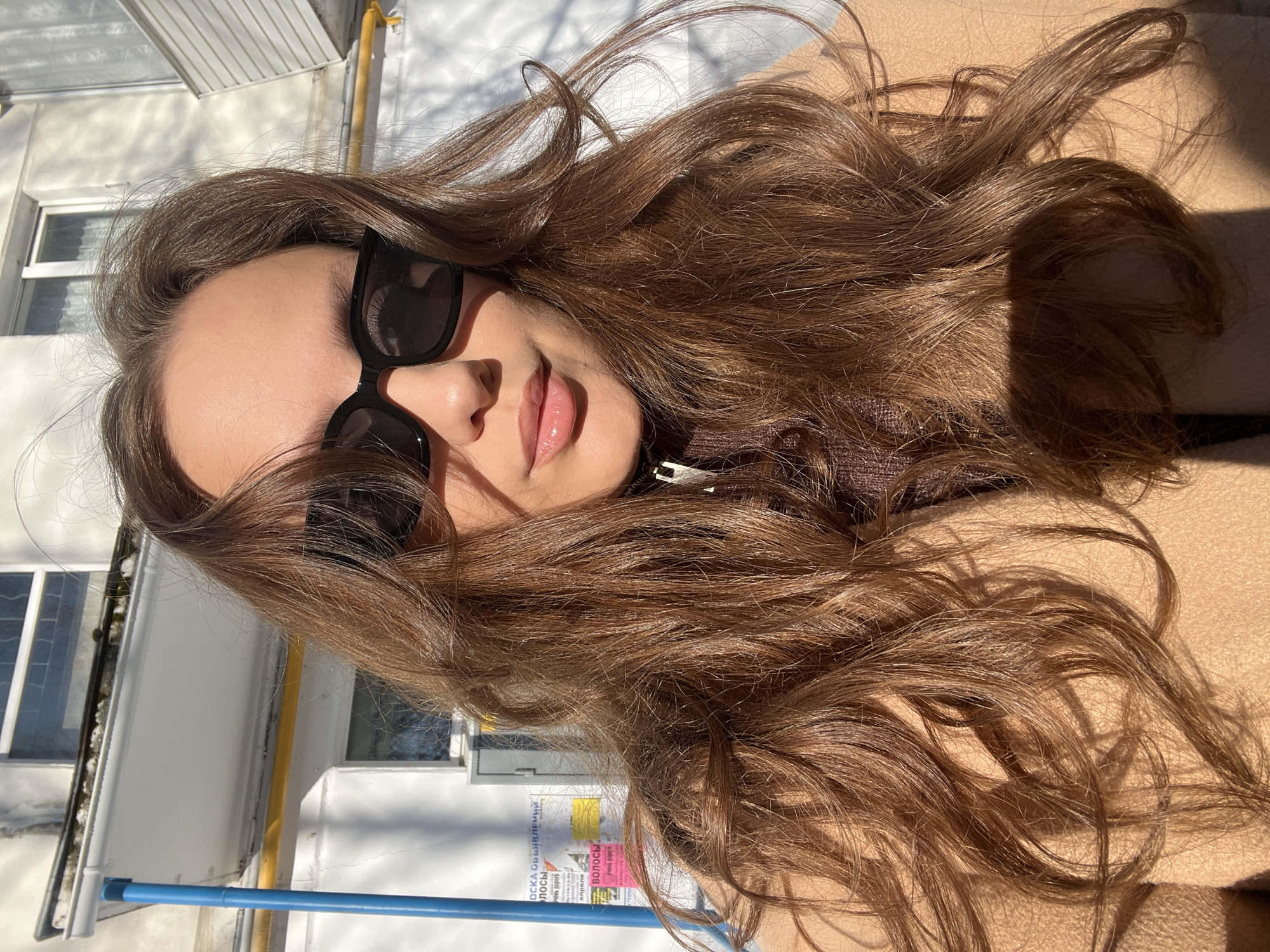 A close-up, eye-level outdoor portrait features an adult female subject positioned centrally in bright sunlight. She has long, wavy, reddish-brown hair cascading over her shoulders and chest. The subject wears black, thick-framed sunglasses and a light brown overcoat, revealing a dark brown, possibly ribbed, collared top with a visible zipper at the neckline. Her lips are visible and appear glossy. Strong direct sunlight illuminates her hair and face, casting defined shadows, notably from her hair onto her coat.
The background consists of a light-colored building, likely residential, with visible window frames. To the left of the subject, a prominent vertical blue pipe runs along the building's exterior, with a yellow horizontal pipe visible below it. Several sheets of paper, acting as posted advertisements or notices with Cyrillic text, are affixed to the building's surface in the upper left background, one of which contains the word "ВОЛОСЫ" (hair). The scene is set on a clear, sunny day in the city of Chaikovsky, Russia.