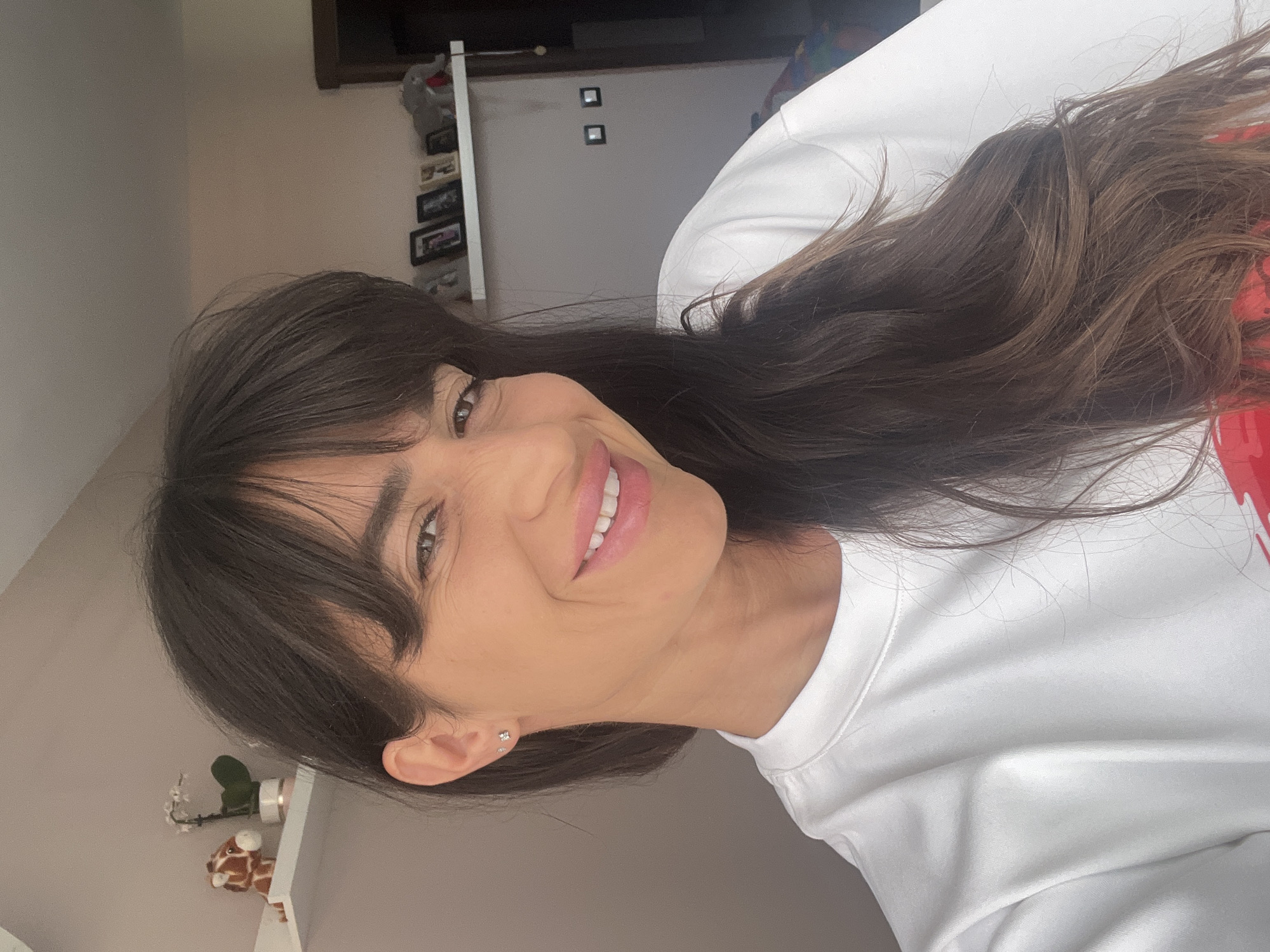 The original image is presented sideways, featuring a close-up portrait of a smiling young woman. She has long, dark brown hair styled with bangs that frame her face. Her head is tilted slightly, and she is smiling broadly, revealing her teeth, with her dark eyes looking directly into the camera. She wears small, subtle stud earrings and a plain white long-sleeved top, with a faint hint of red visible on the right side of her shirt, possibly part of a design.
The background suggests an indoor, domestic setting with light-colored walls. On a white shelf, positioned to the lower left of her face (when viewed upright), a small plant (possibly an orchid) and a spotted giraffe figurine are visible. Further back, on a wall, there are multiple framed pictures or decorative items arranged vertically. Two additional small, square, dark-framed objects are also visible on another section of the wall. The overall lighting is soft and even, illuminating her face and highlighting her pleasant, cheerful expression. No visible text is present in the image.