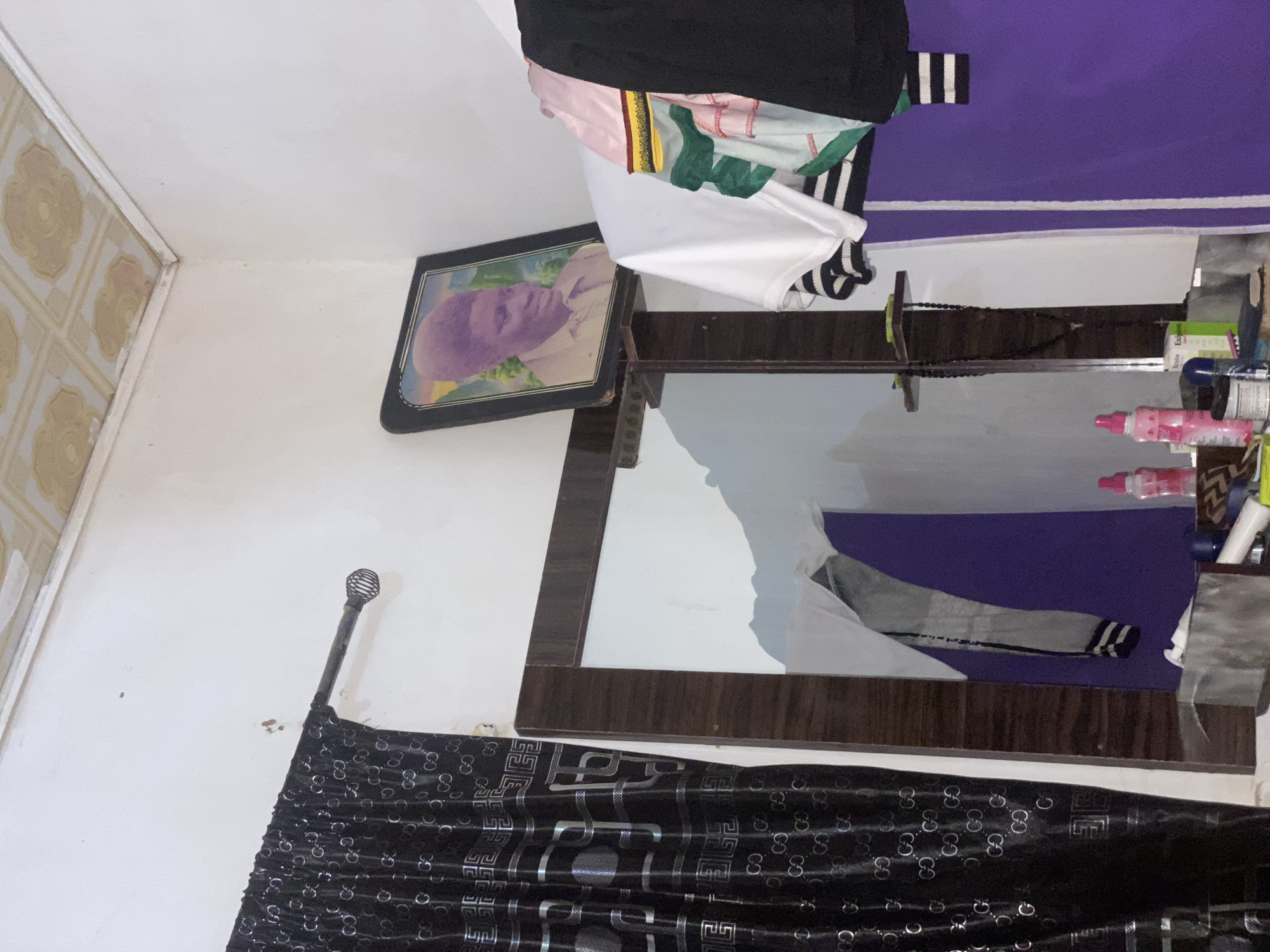 The image depicts a corner of a room, likely a bedroom or dressing area, within a residential setting in Jalingo, Nigeria. The walls are painted a light color, and the ceiling features a decorative tiled pattern, showing some signs of wear.
On the left, a black curtain with a repeating "GG" logo pattern hangs from a dark rod with a decorative finial. To the right of the curtain is a tall mirror with a dark wooden frame. The mirror reflects parts of the room, including a purple-clad wall and what appears to be hanging clothing.
Above the mirror, on the wall, is a framed portrait of a young Black male with an afro hairstyle, wearing a collared shirt. The portrait has a dark frame.
To the right of the portrait, several items of clothing are draped over a railing or shelf. These include a white t-shirt, a t-shirt with horizontal stripes, and a garment with green and red stripes. A black garment is also visible.
On a surface below the mirror and to the right, various items are placed, including two pink spray bottles, a blue-capped container, a white tube, and other unidentifiable objects, suggesting a vanity or dressing area. A dark string of beads with a cross pendant hangs from a small shelf mounted on the mirror frame. The overall impression is of a lived-in space with personal items.