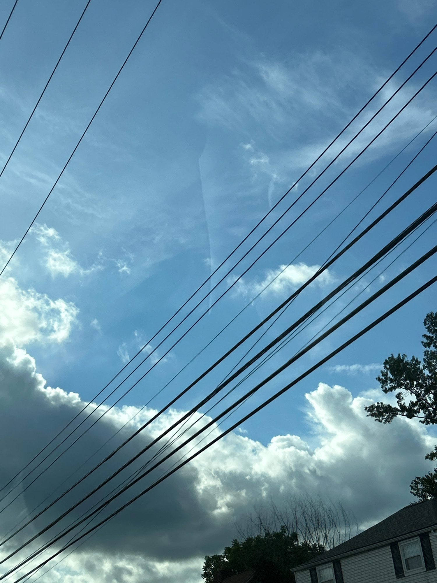 The image shows a view of the sky with overhead power lines in the foreground, taken from a street level perspective. Multiple dark power lines stretch diagonally across the frame, from the bottom left to the top right. The sky is a bright blue, with scattered white and grey clouds. Some of the clouds are large and fluffy, while others are wispy and spread out. On the far right of the image, a portion of a green leafy tree is visible, and below that, the roof and siding of a house can be seen. The house appears to be a light color with dark shutters around the windows. There are no people or animals visible in the image. The scene suggests an outdoor, possibly suburban or residential, environment under a partly cloudy sky. The image was captured in Pittsburgh, United States of America.