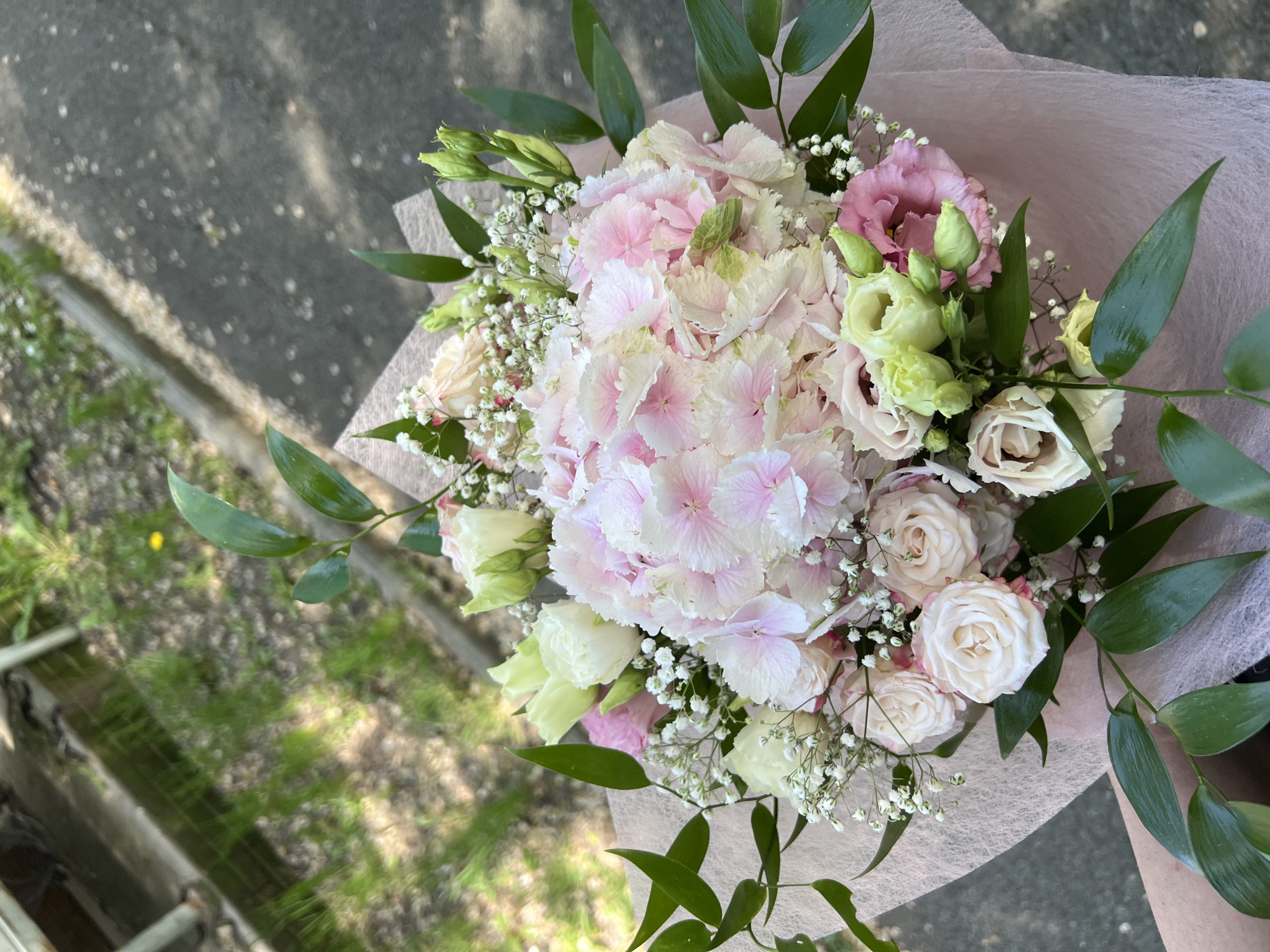 A bouquet of pink and white flowers, wrapped in pink tissue paper, is held in front of an urban scene. The bouquet features hydrangeas, roses, and baby's breath. Green foliage surrounds the flowers. The background includes asphalt, grass, and a weathered concrete structure. The location is Burgas, Bulgaria.
