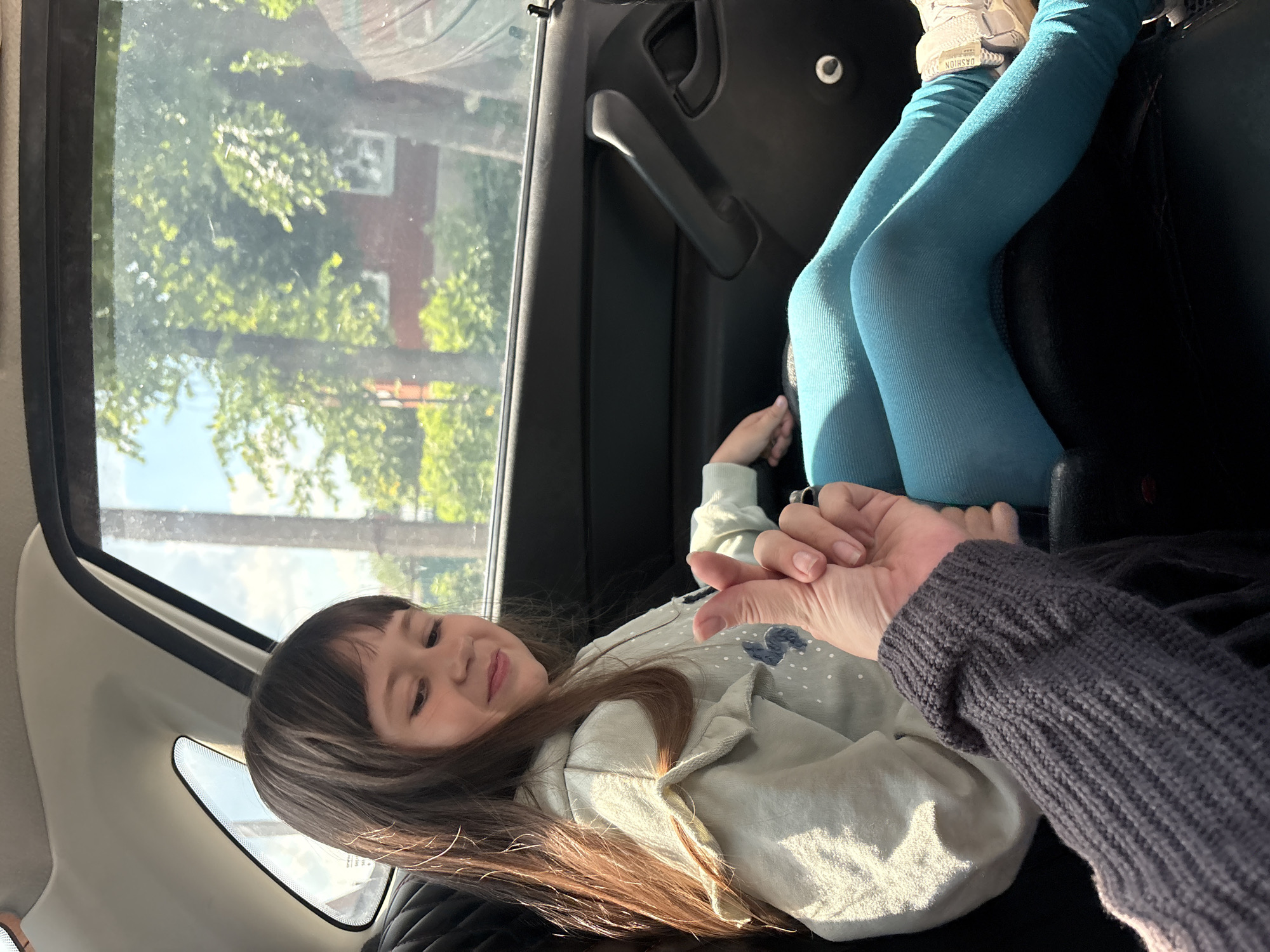 In an interior shot taken from a low angle within a vehicle, a young girl with long, dark brown hair and bangs is seated on the back seat. She is wearing a light blue hooded sweatshirt with small white polka dots and embroidered blue rabbits. She is also wearing teal-colored leggings and light-colored sneakers. Her head is tilted to her right, and she is smiling with her eyes closed. Her right hand is near her chest, and her left hand is obscured. A person's hand, wearing a dark gray ribbed sweater, is extended towards the girl, with their thumb and index finger making a small heart shape gesture. The girl appears to be looking at this gesture. The background shows a car's interior, with the door handle visible, and through the window, lush green trees and a red-bricked building are visible under a clear blue sky. The scene is likely on a street or road within the city of Chaikovsky, Russia, given the presence of trees and residential buildings, common in such urban environments.