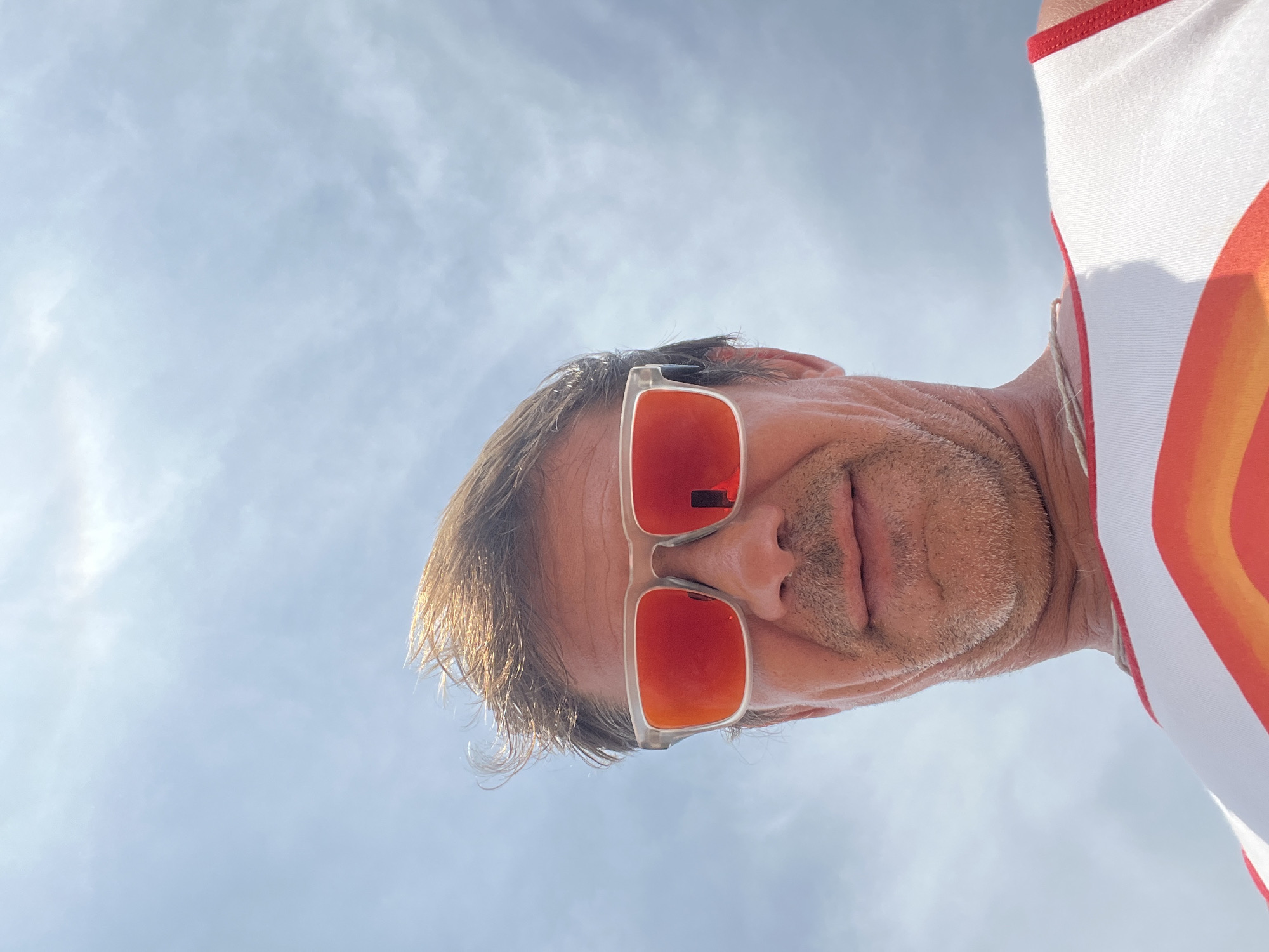 A low-angle, close-up, outdoor, selfie shot shows a man wearing sunglasses with orange lenses and a white tank top with a rainbow design. The man's hair is windswept, and he has stubble on his chin. He appears to be outdoors on a cloudy day, with a bright sky visible in the background.