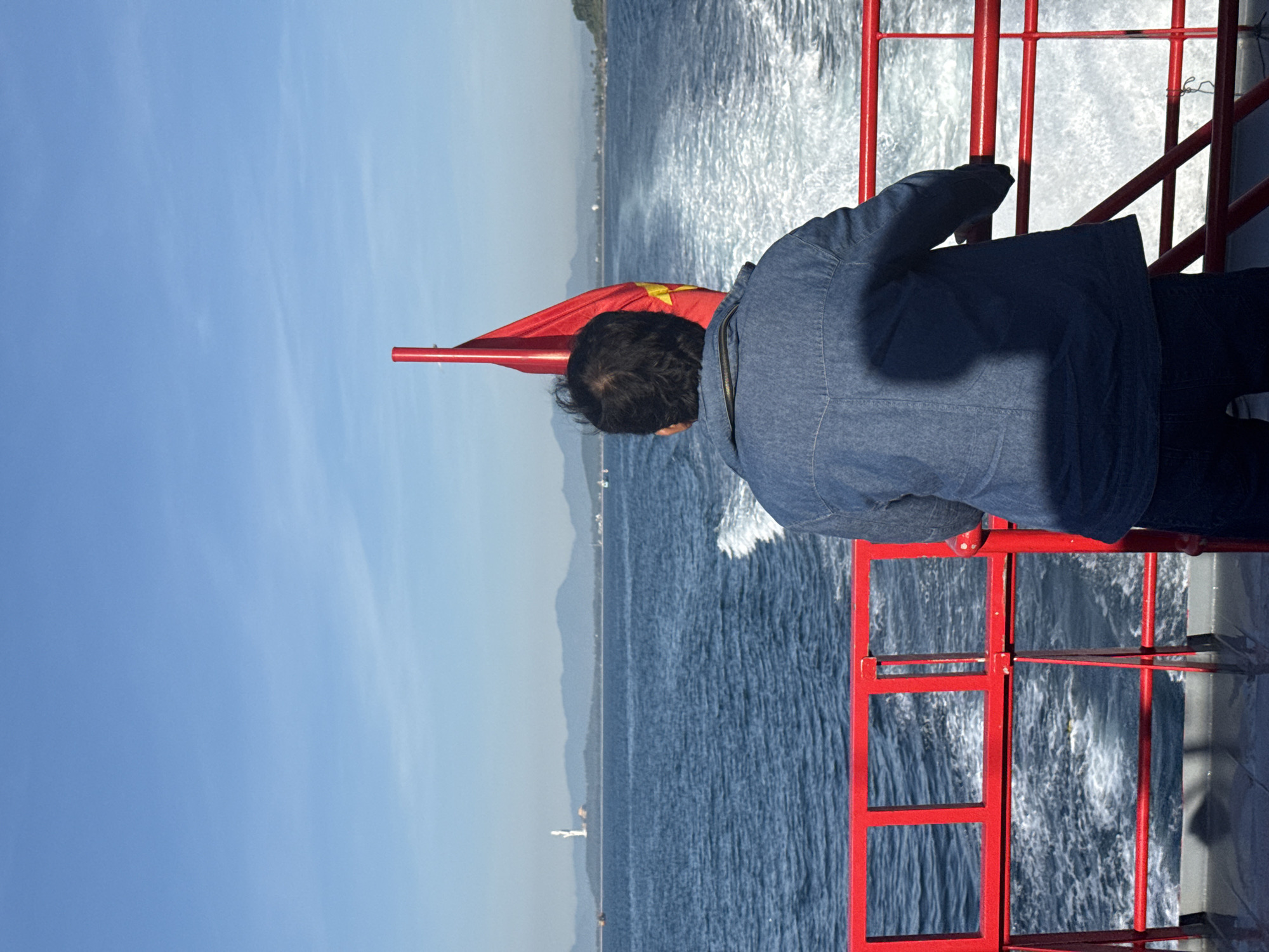 A man in a denim jacket stands on the deck of a red-railed ferry, looking out at the ocean. A Vietnamese flag flies from a pole behind him. The ferry is moving, creating a white wake in the blue water. In the distance, a coastline with mountains and buildings is visible. A tall, white statue stands on a hill on the distant shore. The sky is clear and blue. The scene is a view from the ferry as it travels on the sea near Tịnh Khê, Vietnam.