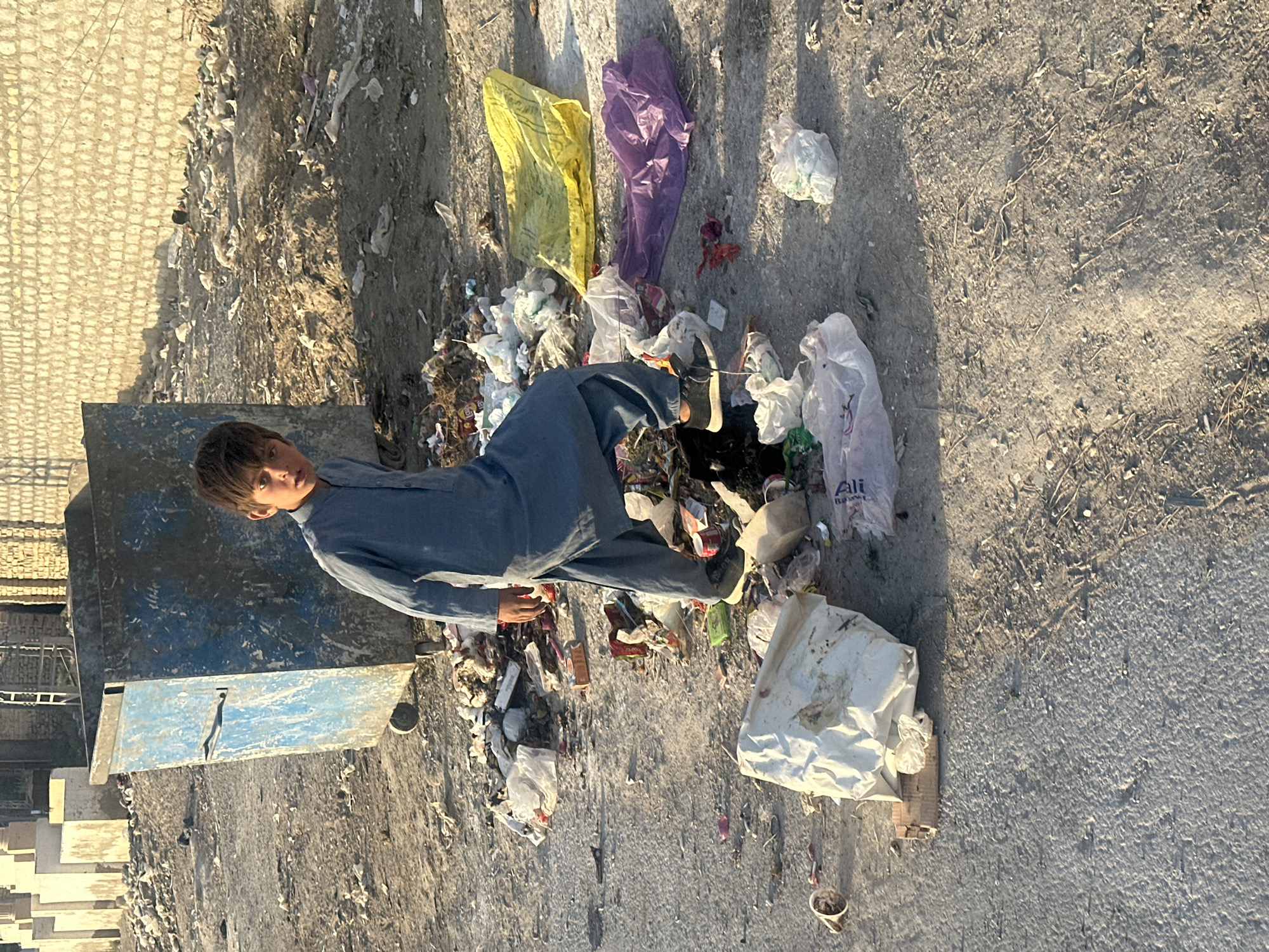 A young boy in a blue outfit stands amidst trash and litter in Quetta City Tehsil, Pakistan. The trash includes various plastic bags, food wrappers, and other debris. He is standing near a blue metal dumpster. The area appears to be an unpaved lot or roadside, with evidence of construction or demolition in the background. The lighting indicates it is daytime.