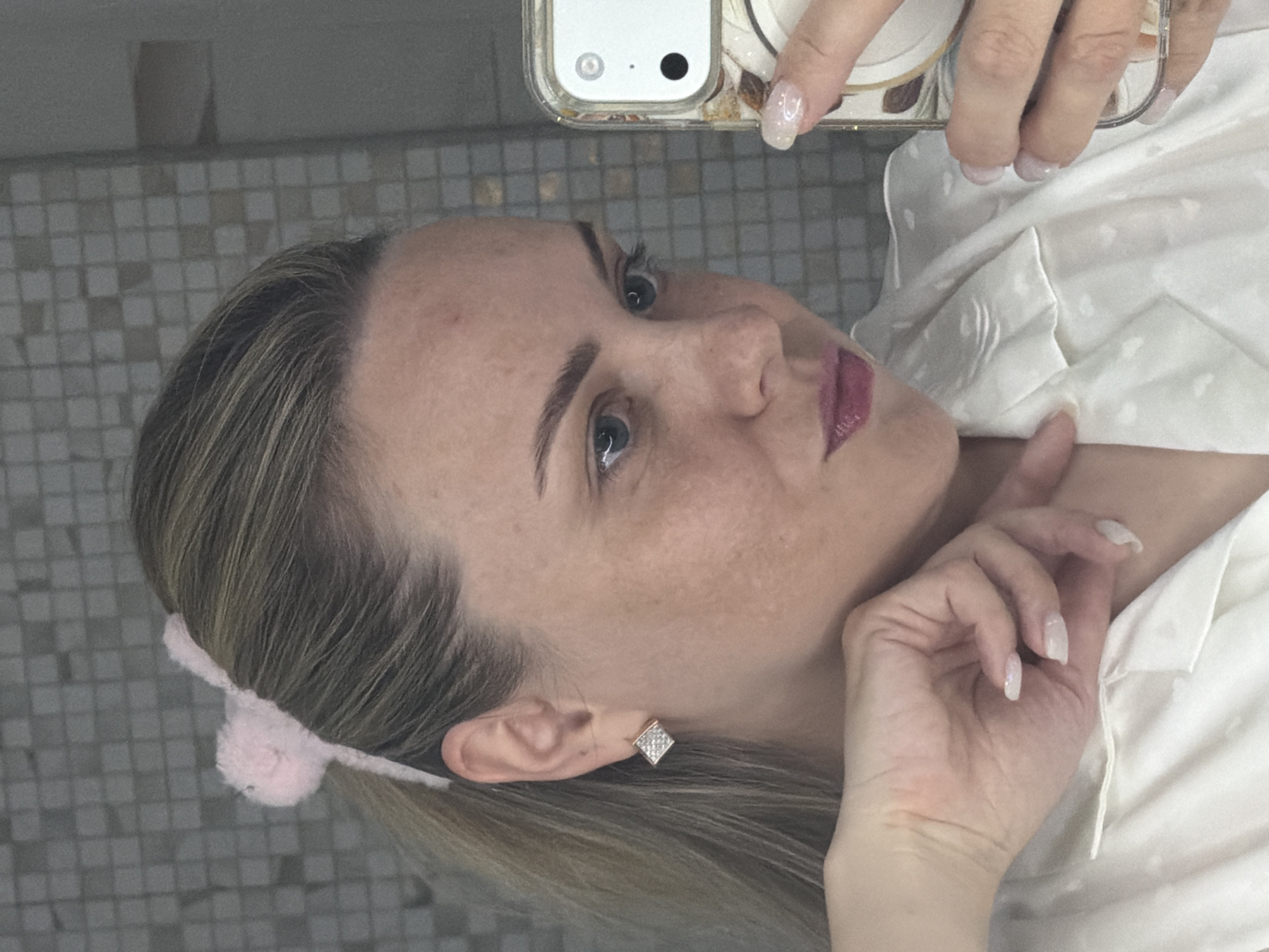 A woman is captured in a close-up mirror selfie, looking directly into the camera with a neutral expression. She has light brown hair with blonde highlights, styled back from her face, revealing a square, studded earring in her right ear. A light pink, fluffy pom-pom hair accessory is visible behind her left ear. Her fair complexion shows some freckles or minor blemishes, and she has well-defined eyebrows and blue eyes. Her lips are adorned with a deep pink or plum-colored lipstick.
She is wearing a light-colored, possibly white or off-white, pajama top or shirt featuring a subtle pattern, possibly small hearts or dots. Her right hand, holding a white smartphone to capture the image, displays long, light-colored artificial nails with a glittery finish. The phone features a distinctive dual-camera module and is encased in a clear cover with decorative elements. Her left hand is casually placed near her chin and neck.
The background consists of a mosaic-tiled wall composed of small, square tiles in shades of gray, white, and beige, suggesting an indoor setting like a bathroom or a dressing area in Votkinsk, Russia. No specific time of day or outdoor weather conditions are discernible, and there is no visible text in the image.