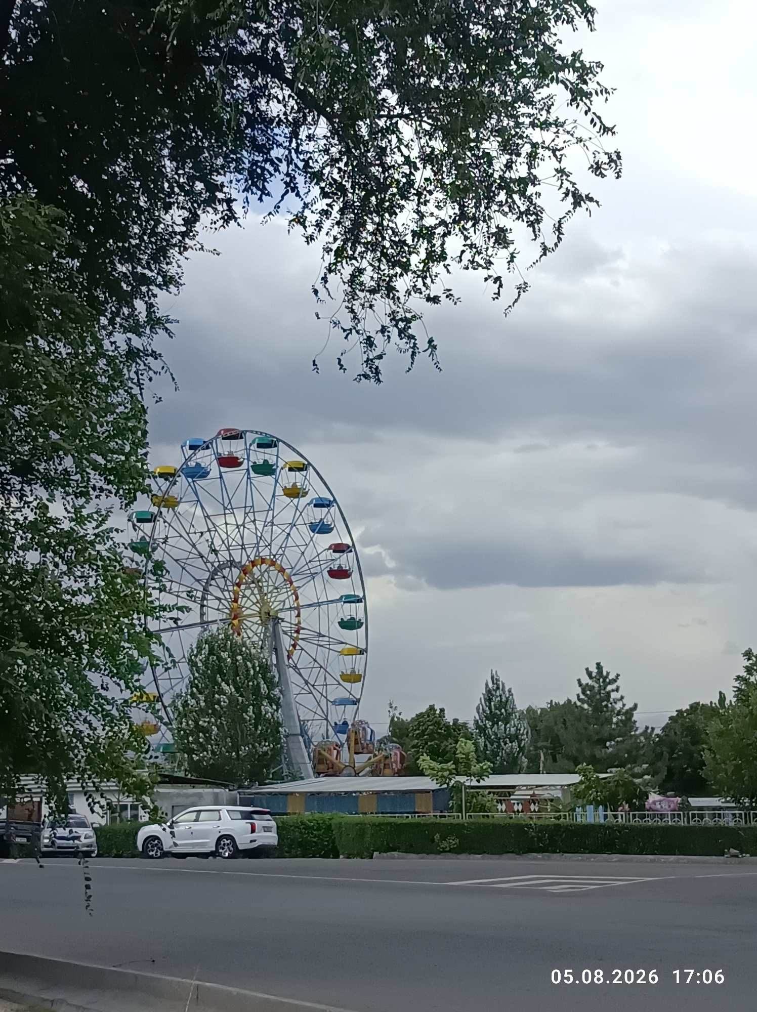 The image shows a Ferris wheel at an amusement park in Tokmok City, Kyrgyzstan. The Ferris wheel has multicolored gondolas and is silhouetted against a cloudy sky. In the foreground, there is a street with two cars parked on the side. Trees line the street, and in the background, there are more trees and buildings. The date and time "05.08.2026 17:06" are visible in the bottom right corner of the image.