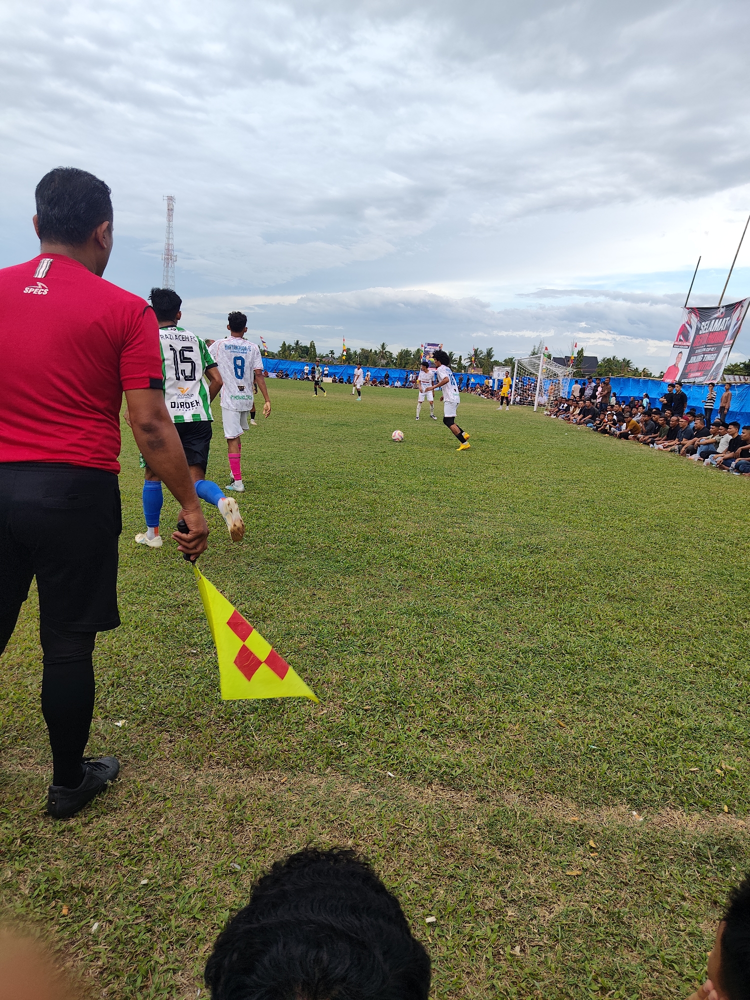 The image depicts a soccer match in progress at a field in Cibrekbaroh, Indonesia. In the foreground, a person in a red shirt and black shorts, likely a coach or official, stands with a yellow and red checkered flag. Several players in soccer uniforms are visible on the green grass field. One player in a green and white striped jersey with the number 15 is running. Another player in a white jersey with the number 8 is also on the field. Spectators are seated along the sidelines, behind a blue barrier. Several banners are displayed in the background. A soccer goal is visible on the right side of the field. The sky is cloudy. The ground is covered in green grass with some patches of dry, brown grass.