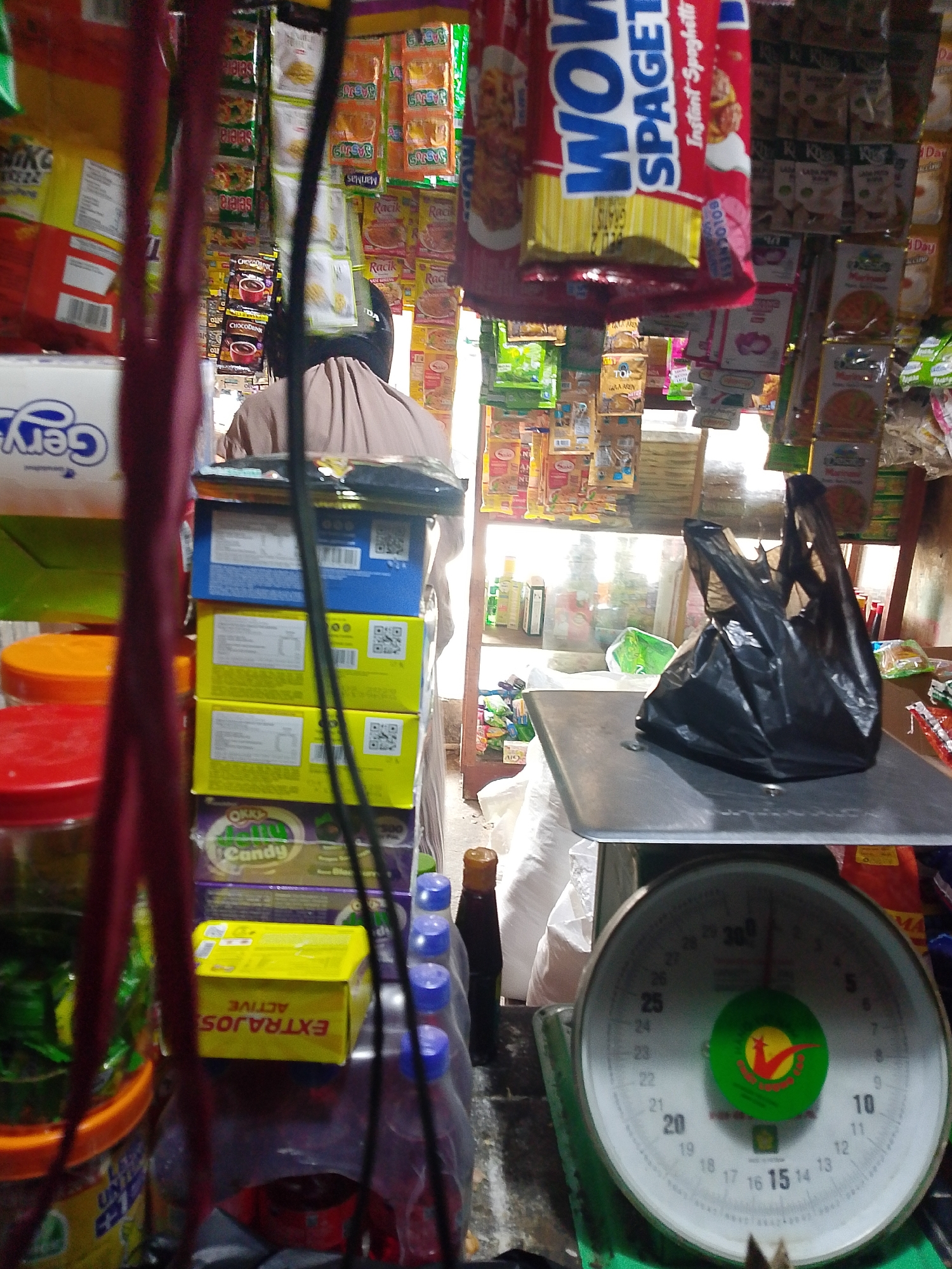 The image shows a close-up of a small convenience store or a market stall in Palembang, Indonesia, with items stacked and displayed. In the foreground, a red strap hangs from above, obscuring part of the view. To the left, several plastic containers with lids are visible, some containing green items. Behind these, there are stacked boxes, one blue and two yellow, with printed text and QR codes.
In the middle ground, a person wearing a light brown garment and a black head covering is partially visible, appearing to be behind a counter or display. To the right of the person, on a metal weighing scale, is a black plastic bag filled with items. The scale has a round dial with numbers indicating weight, and a green circular logo with red and yellow markings.
The background is filled with shelves stocked with various packaged goods, including brightly colored snack bags, boxes, and bottles. The lighting suggests an indoor setting, possibly with natural light filtering in from an opening or window. The overall impression is of a busy retail environment, typical of a small local business in a city like Palembang.