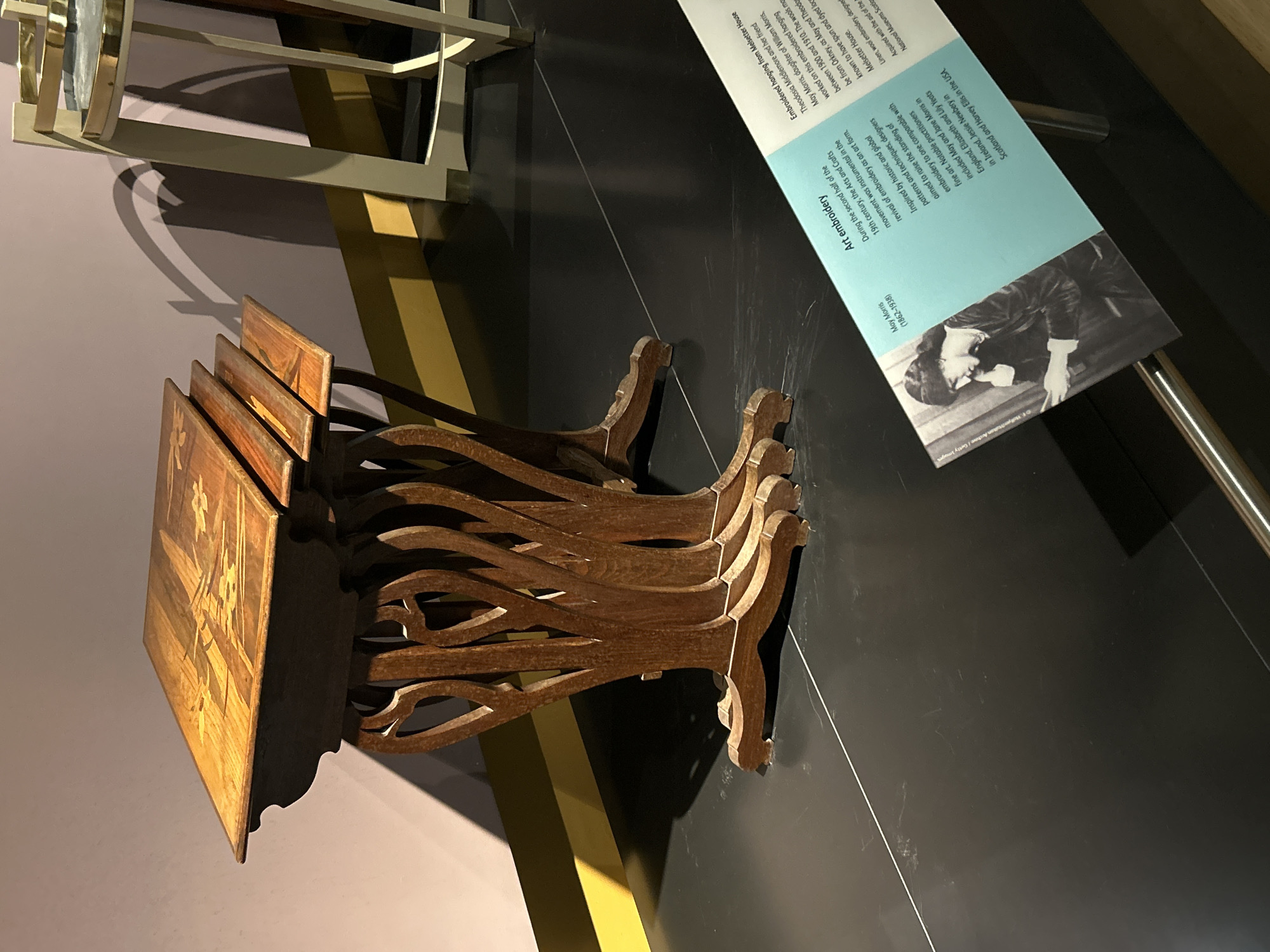 This image shows a display of antique furniture and an explanatory sign within a museum setting, specifically at the National Museums Scotland in the city of Edinburgh, United Kingdom. The primary focus is a set of nesting tables made of dark wood, with intricate inlay designs on their surfaces. The tables are elegantly carved with a motif resembling organic, flowing branches or roots supporting the tabletops.
To the right of the nesting tables, a portion of another display piece is visible, appearing to be a stool or small table with a metal frame and circular top.
In the foreground, a placard with an image and text provides information about "Art embroidery." The image on the placard is a black and white photograph of a woman, identified as May Morris (1862-1938), who is shown in profile, looking downwards, engaged in needlework. The text details the revival of embroidery as an art form during the 19th century, its association with historic design, and notable practitioners. The placard is angled on a metal stand.
The floor of the exhibit space is a dark, reflective surface, with a thin white line running diagonally across it. The background features a wall with a gentle gradient of color, transitioning from a light purple to a darker hue, with a distinctive curved shadow cast upon it, suggesting an external light source or architectural element. A yellow stripe runs along the base of the wall. The overall impression is of a well-lit museum exhibit designed to showcase historical decorative arts.