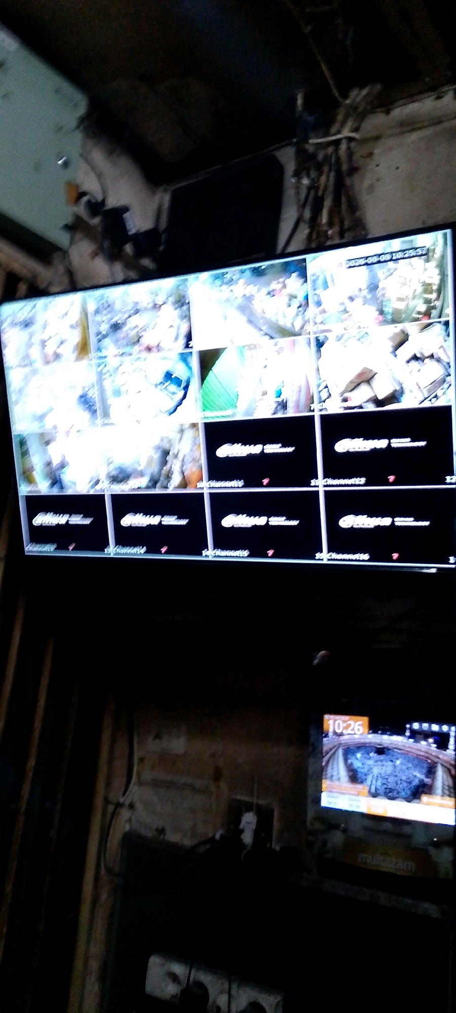 A surveillance system display shows multiple camera feeds, with one camera feed showing the date "2026-09-09 10:25:57". Another smaller screen in the lower right corner shows a single camera feed with the time "10:26". The overall environment appears to be a cluttered and dimly lit interior space, possibly a storage area or workshop. The image is taken from a low angle, looking up at the screens and the ceiling. The location is identified as Bandung City, Indonesia.